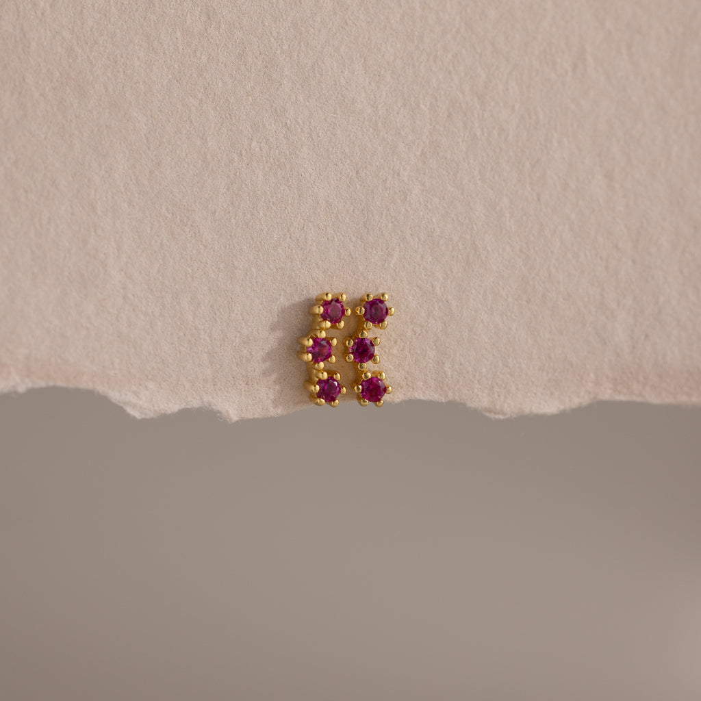 The Ruby Cluster Flatback Studs feature a small gold cluster design with six pink gemstones, shown on a textured beige surface.