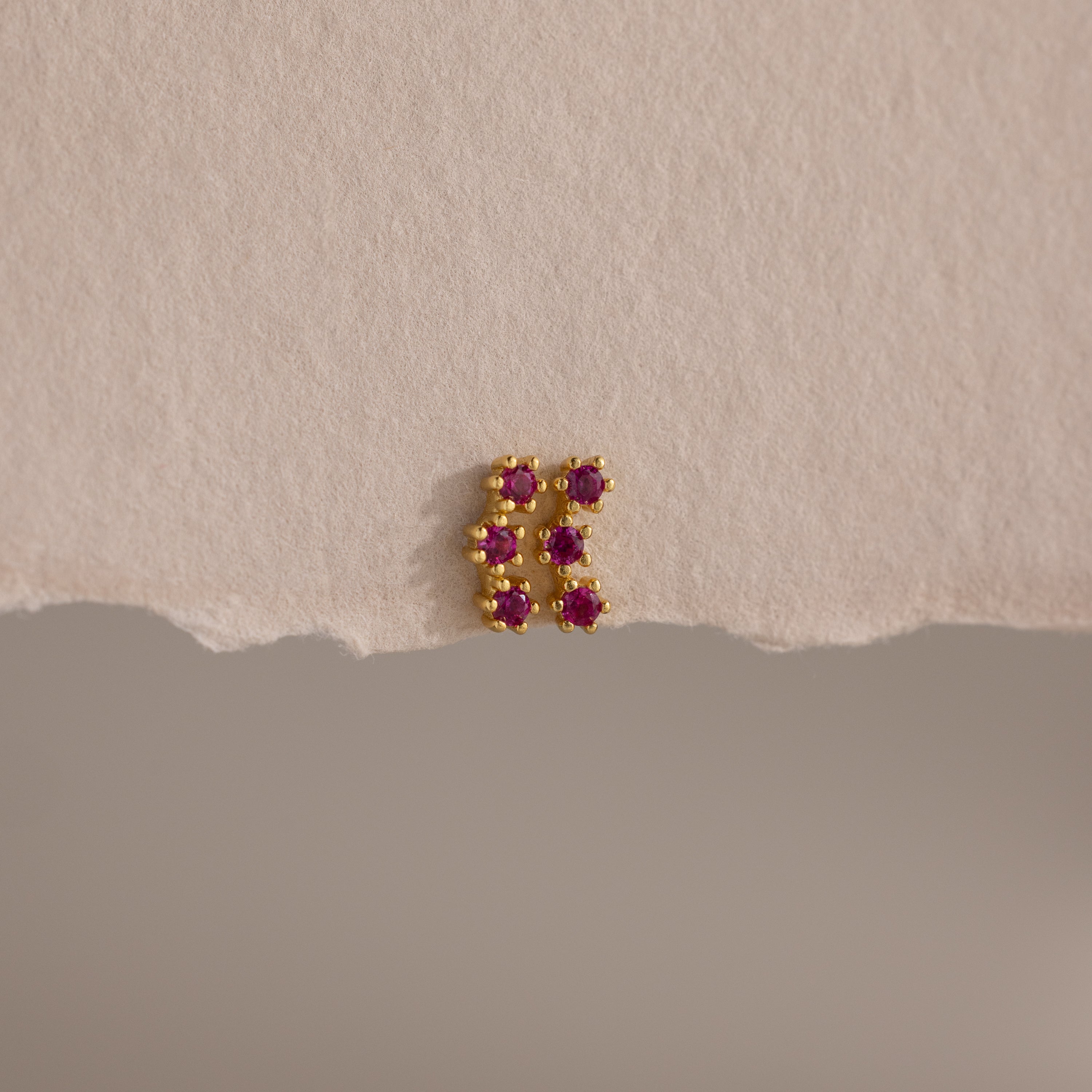 The Ruby Cluster Flatback Studs feature a small gold cluster design with six pink gemstones, shown on a textured beige surface.