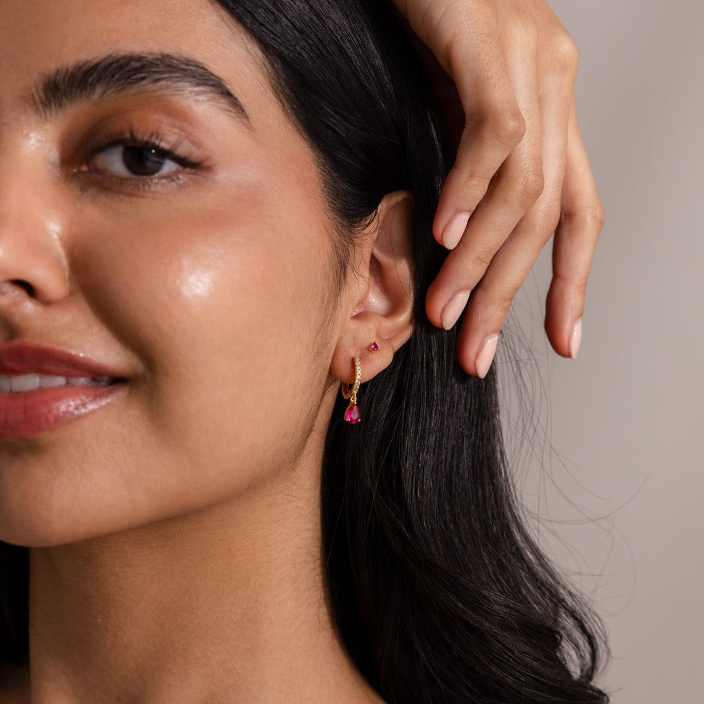 Woman smiling, showing off a gold hoop earring with a pink gemstone and matching pink Teardrop Birthstone Flatback Studs—ideal for fans of Birthstone Jewelry.