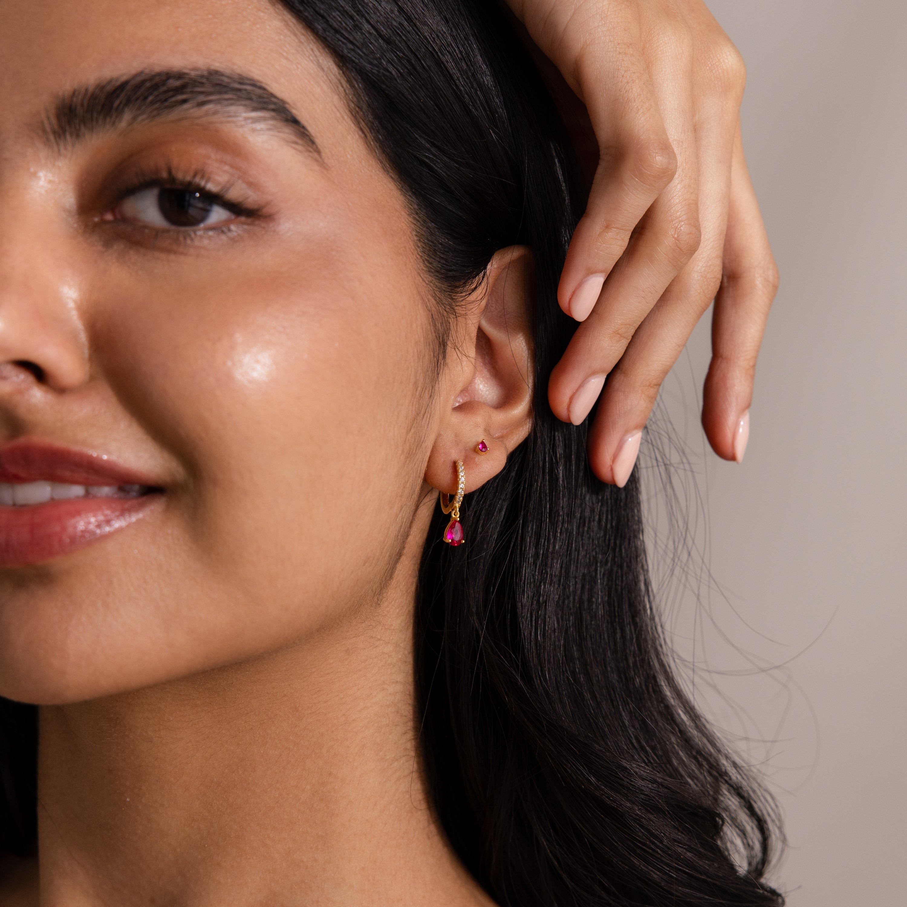 Woman smiling, showing off a gold hoop earring with a pink gemstone and matching pink Teardrop Birthstone Flatback Studs—ideal for fans of Birthstone Jewelry.