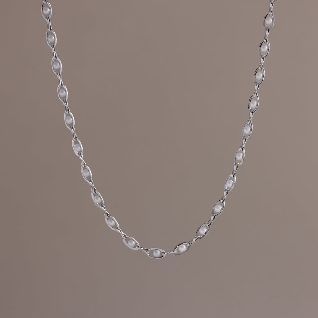 Close-up of a delicate silver chain necklace with oval pearl links, suspended against a neutral backdrop.