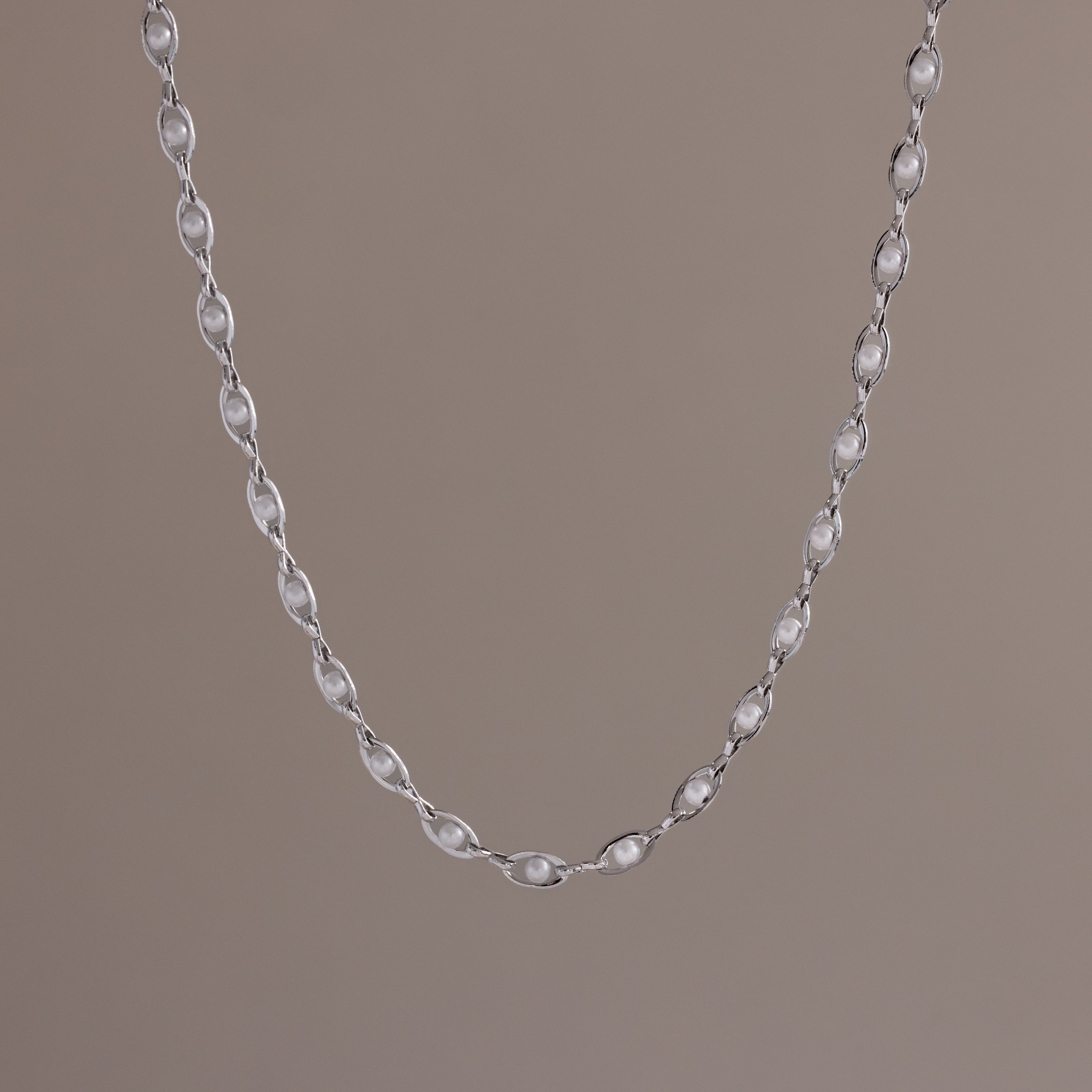 Close-up of a delicate silver chain necklace with oval pearl links, suspended against a neutral backdrop.