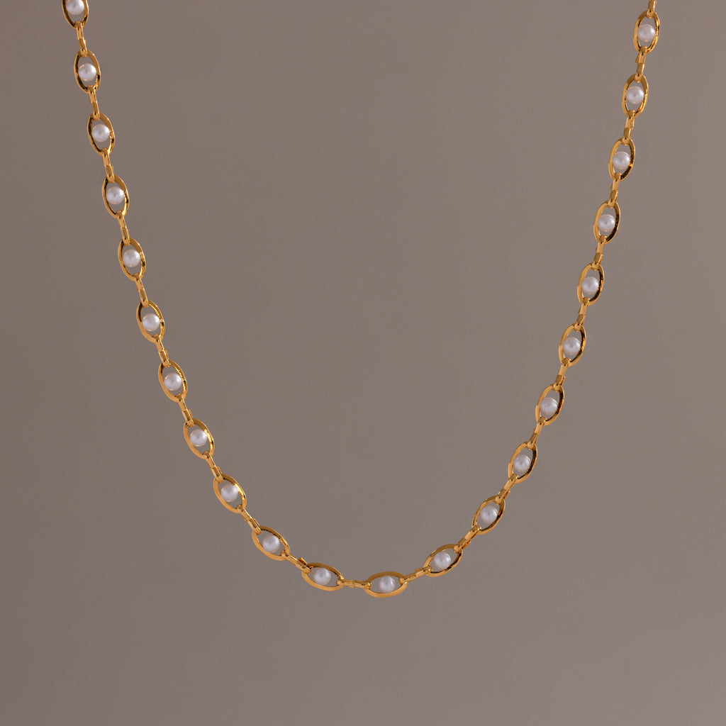 Close-up of a delicate gold chain necklace with oval pearl links, suspended against a neutral backdrop.
