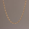 Close-up of a delicate gold chain necklace with oval pearl links, suspended against a neutral backdrop.