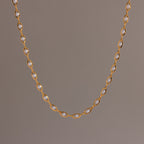 Close-up of a delicate gold chain necklace with oval pearl links, suspended against a neutral backdrop.