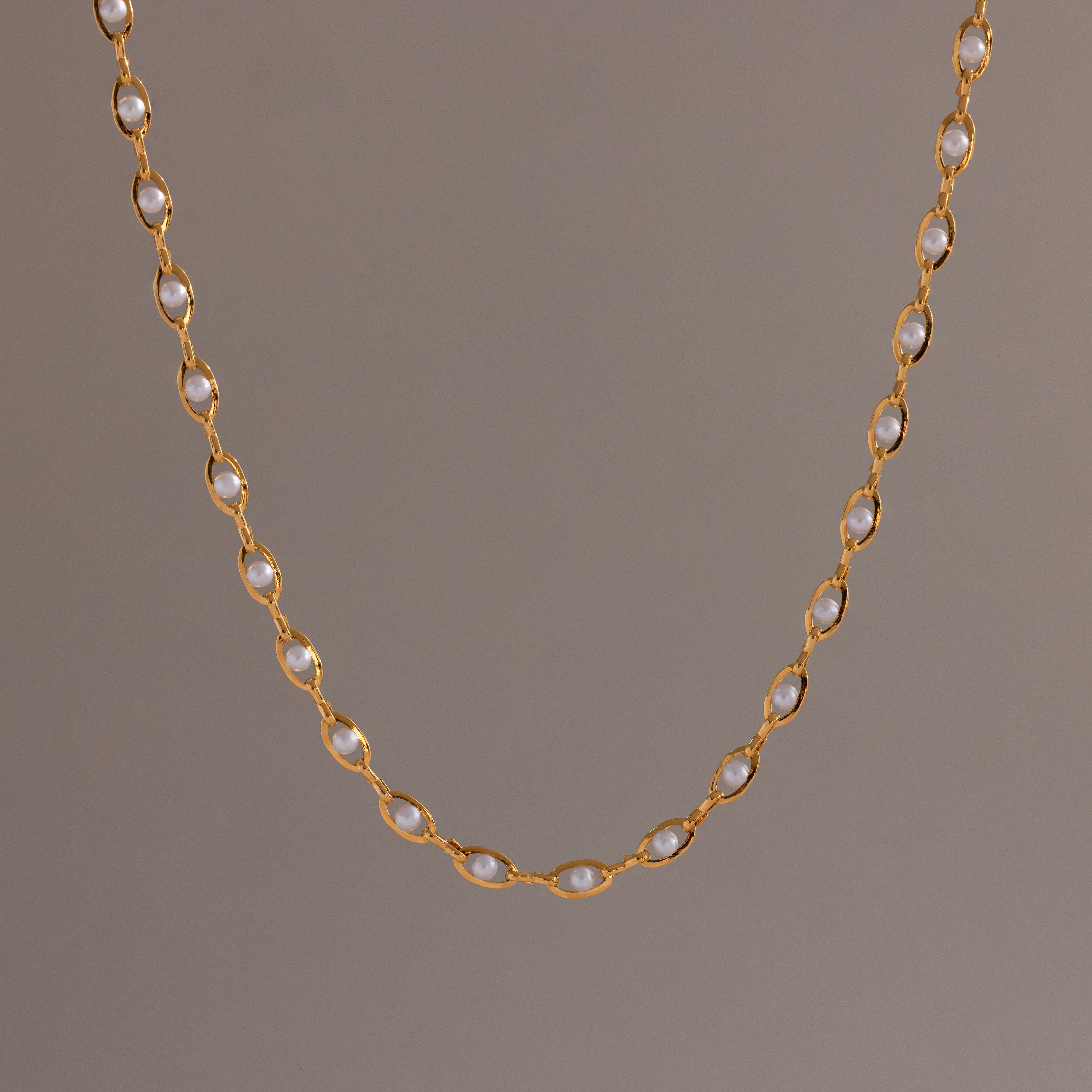 Close-up of a delicate gold chain necklace with oval pearl links, suspended against a neutral backdrop.