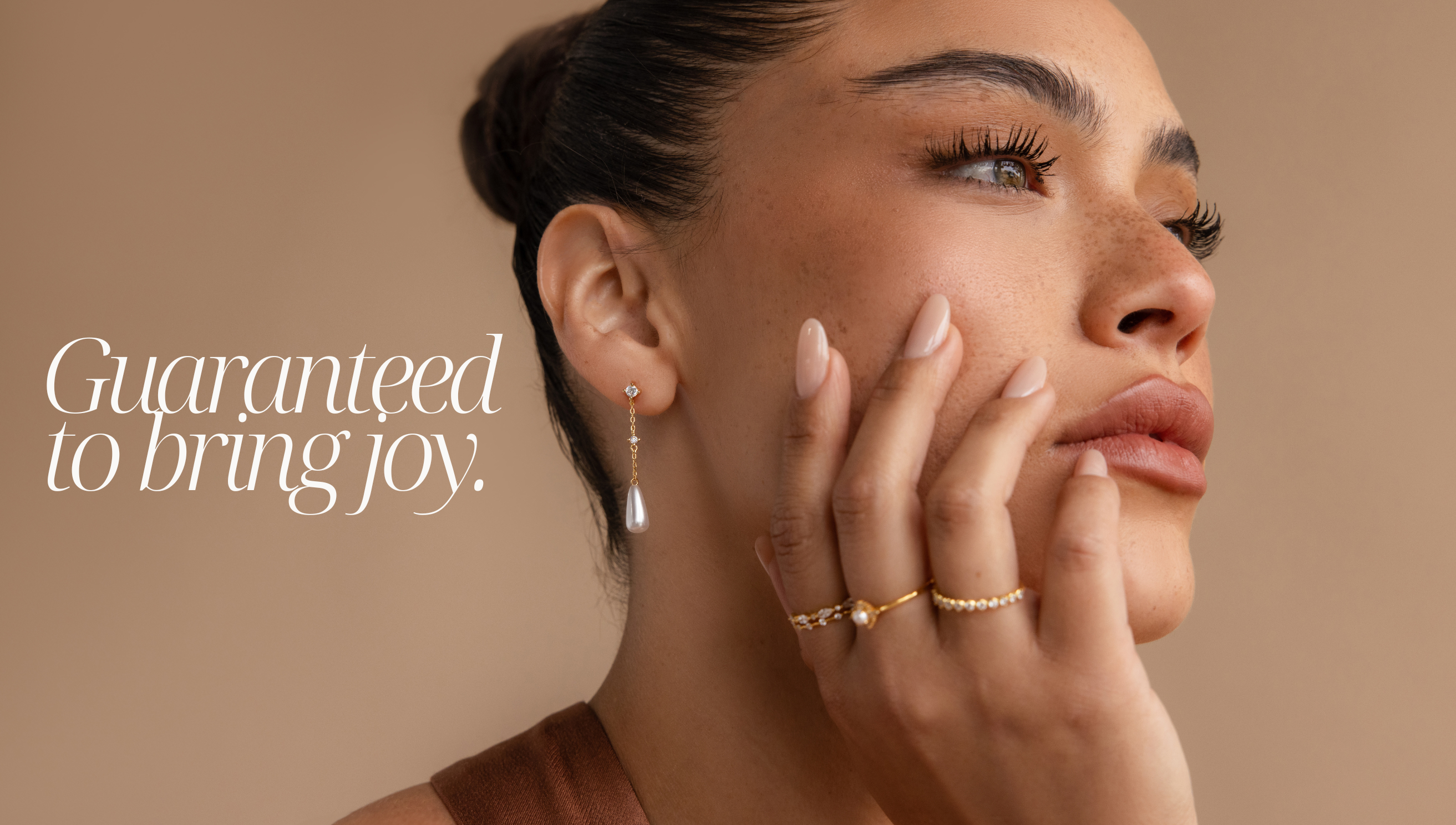 Best Sellers feature photo with the text “guaranteed to bring joy” featuring a jewelry model wearing gold dangling pearl diamond studs on her ear, and a diamond and pearl gold ring stack on her index, middle and ring fingers.