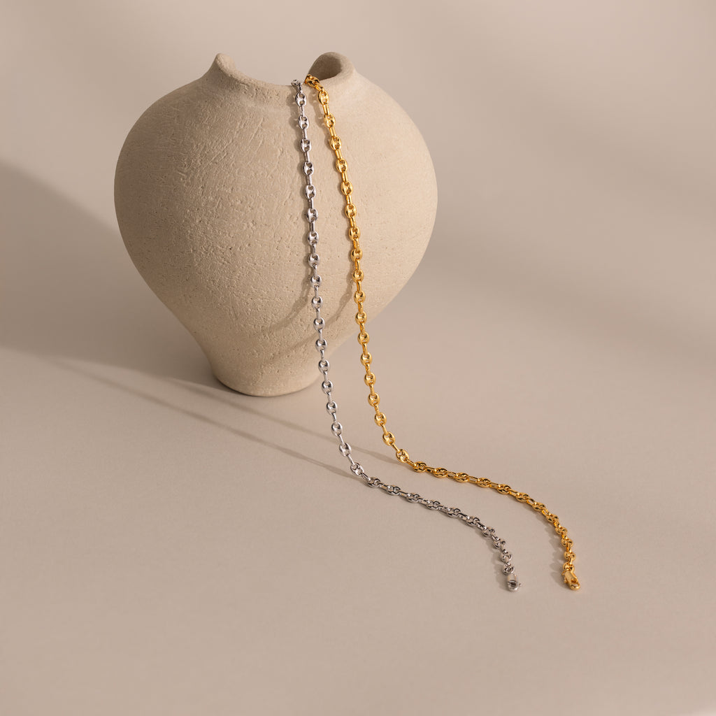 Gold and silver mariner chain necklaces with polished oval links, draped side by side over a textured beige ceramic vase.