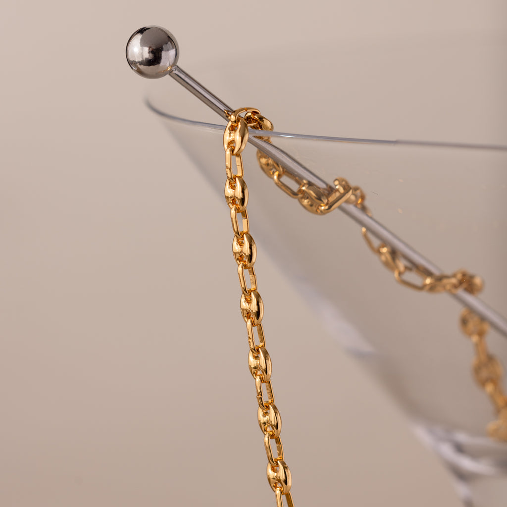 Close-up of a gold mariner chain necklace with polished oval links, elegantly draped over the rim of a clear martini glass with a silver stir stick.