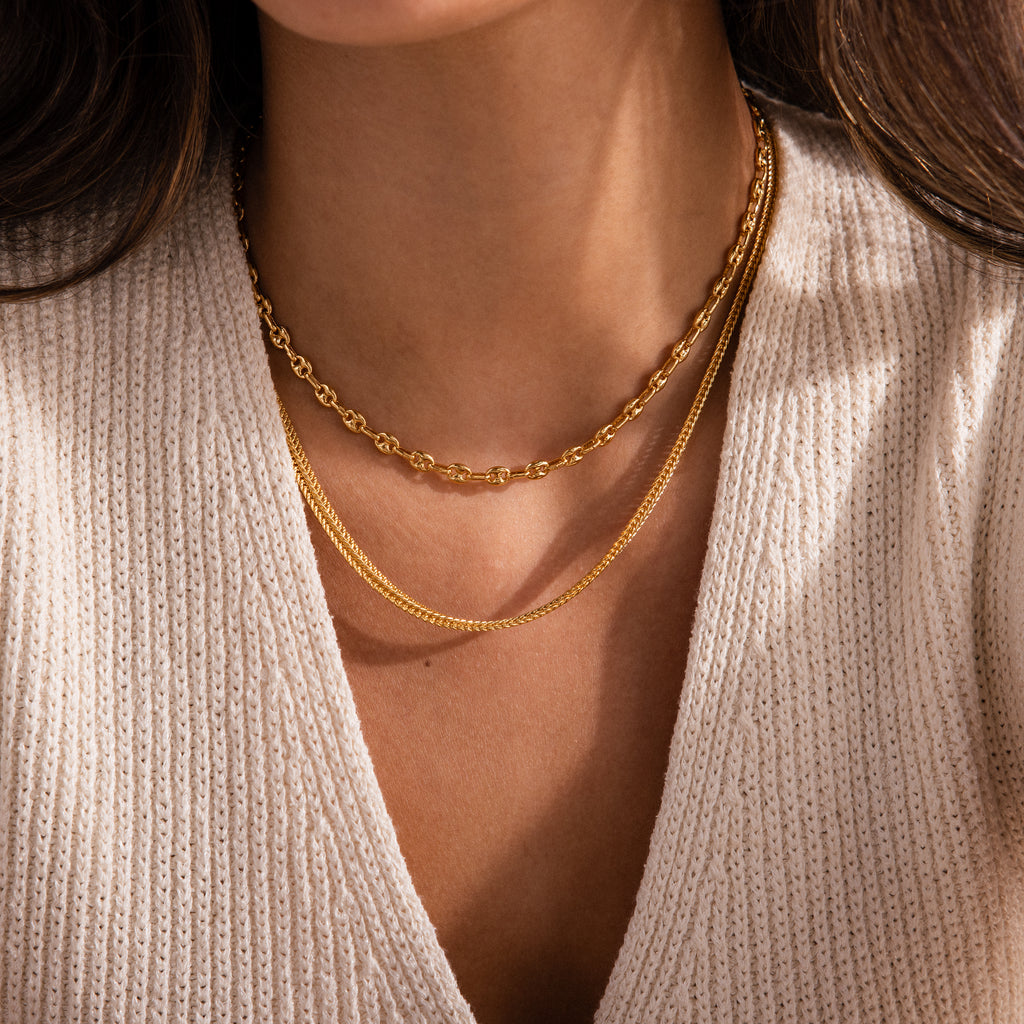 Model wearing layered gold necklaces, including a mariner chain necklace and a thin mesh chain.