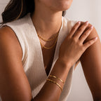 Model wearing layered gold jewelry, including a mariner chain necklace, a thin mesh chain necklace, a mini mariner chain bracelet, and a gold herringbone bracelet, styled with a sleeveless knit top.