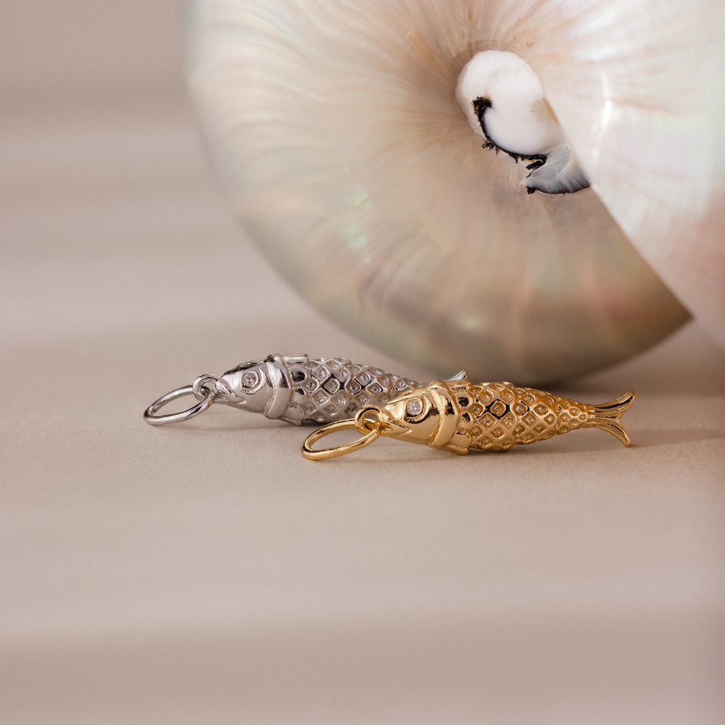 Gold and silver fish charm pendants with textured scale details, displayed beside a seashell.