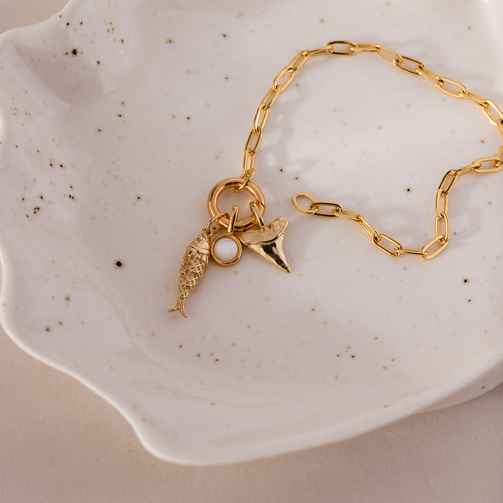 Gold chain bracelet with three ocean-inspired charms: a textured fish, a round white accent, and a shark tooth pendant.