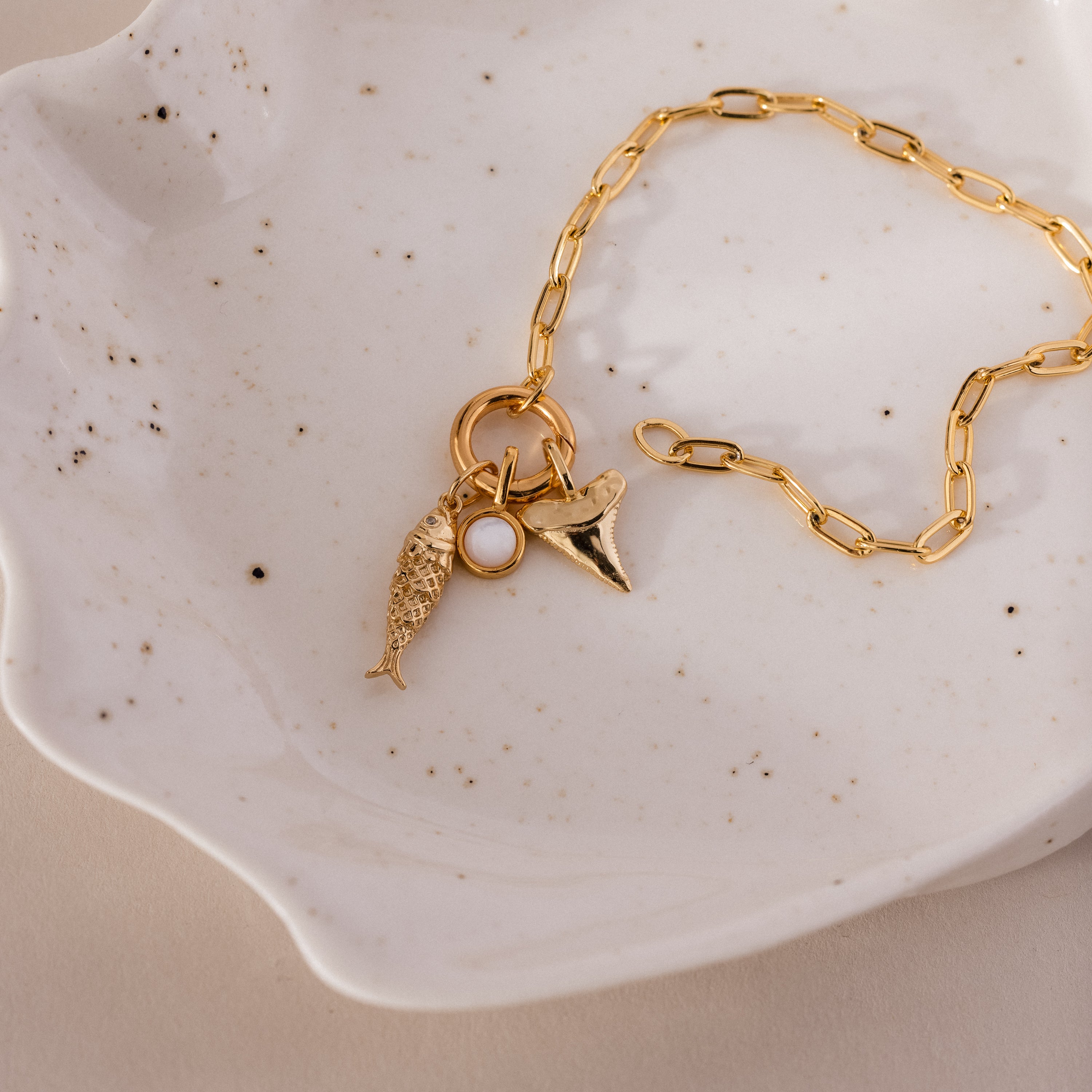 Gold chain bracelet with three ocean-inspired charms: a textured fish, a round white accent, and a shark tooth pendant.