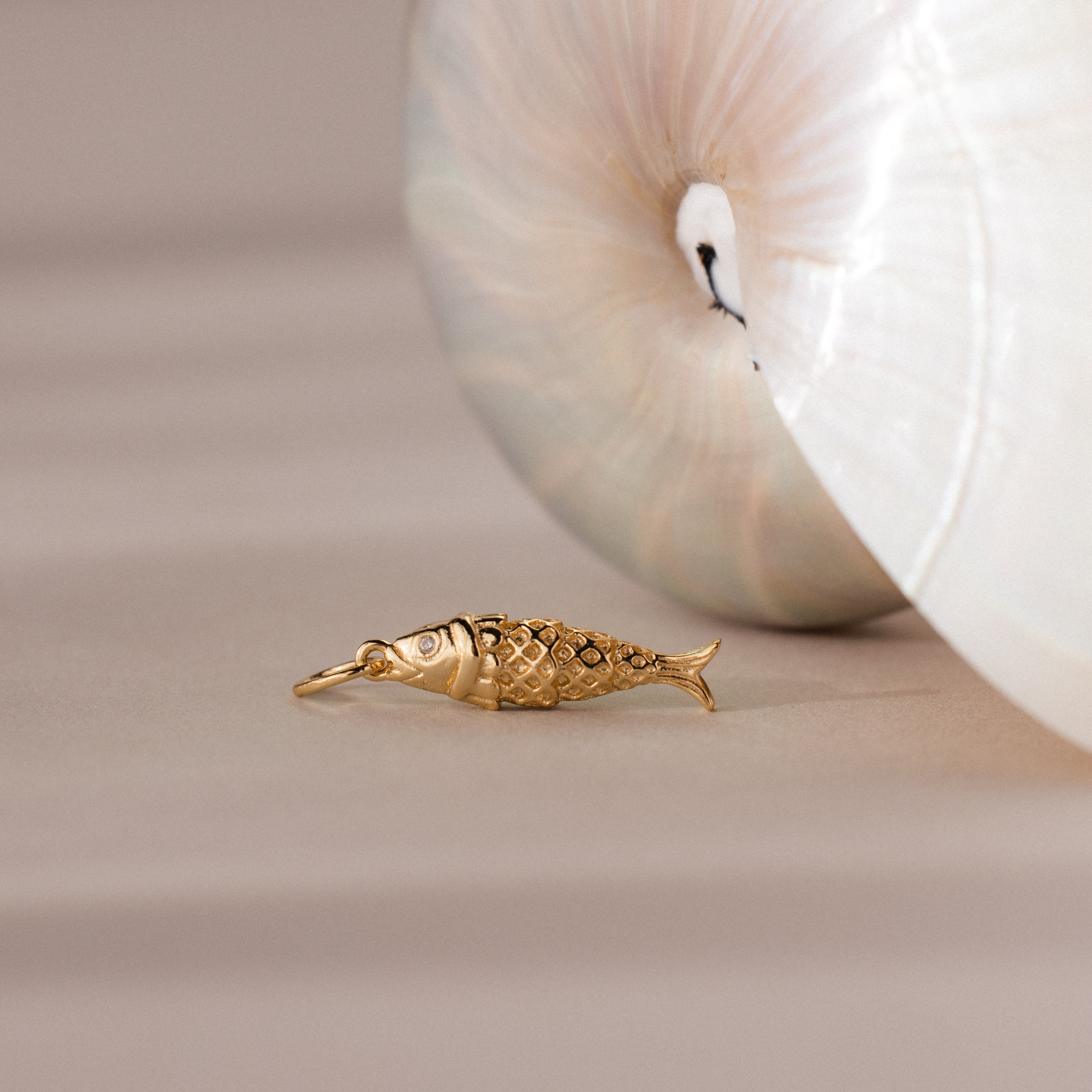 Fish Charm in 18K Gold laying next to a shell