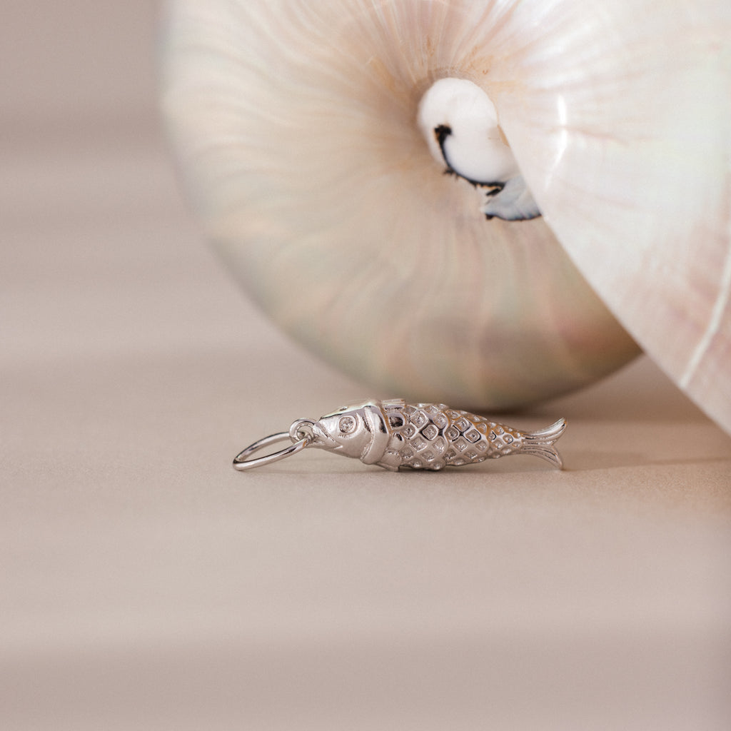 Silver fish charm with detailed scale texture, styled beside a pearlescent seashell.