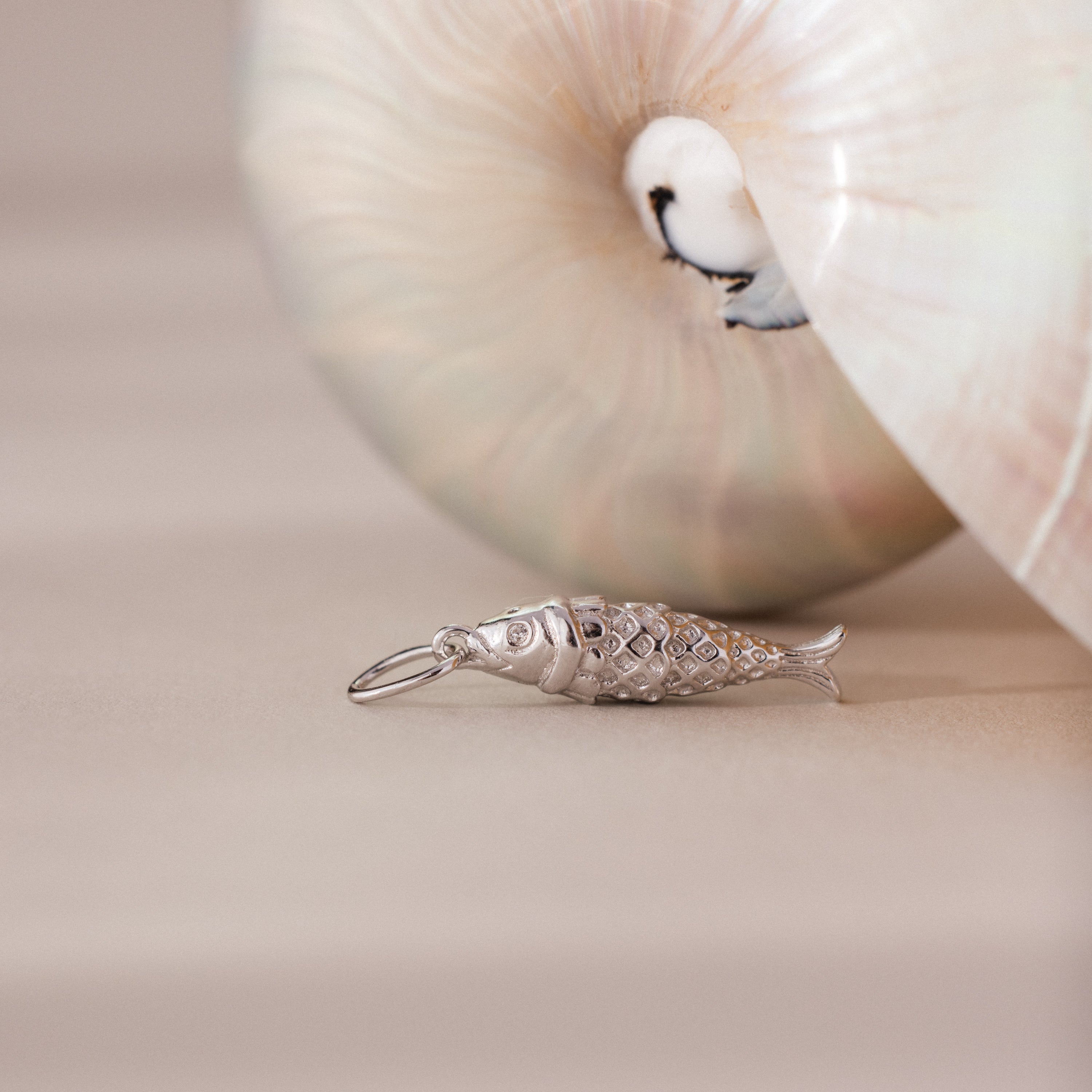 Silver fish charm with detailed scale texture, styled beside a pearlescent seashell.