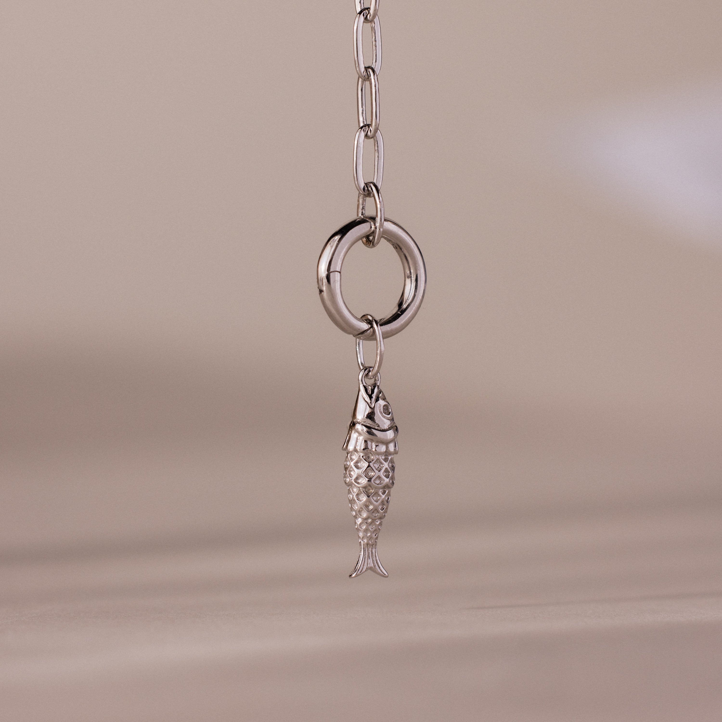 Silver fish charm pendant with intricate scale detailing, hanging from a polished circular link on a chain.