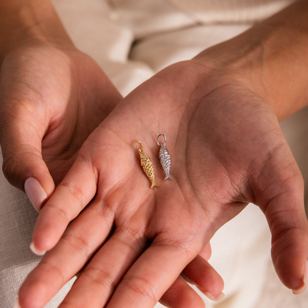 Close-up of hands holding two textured fish charms, one in gold and one in silver, showcasing detailed scale design.