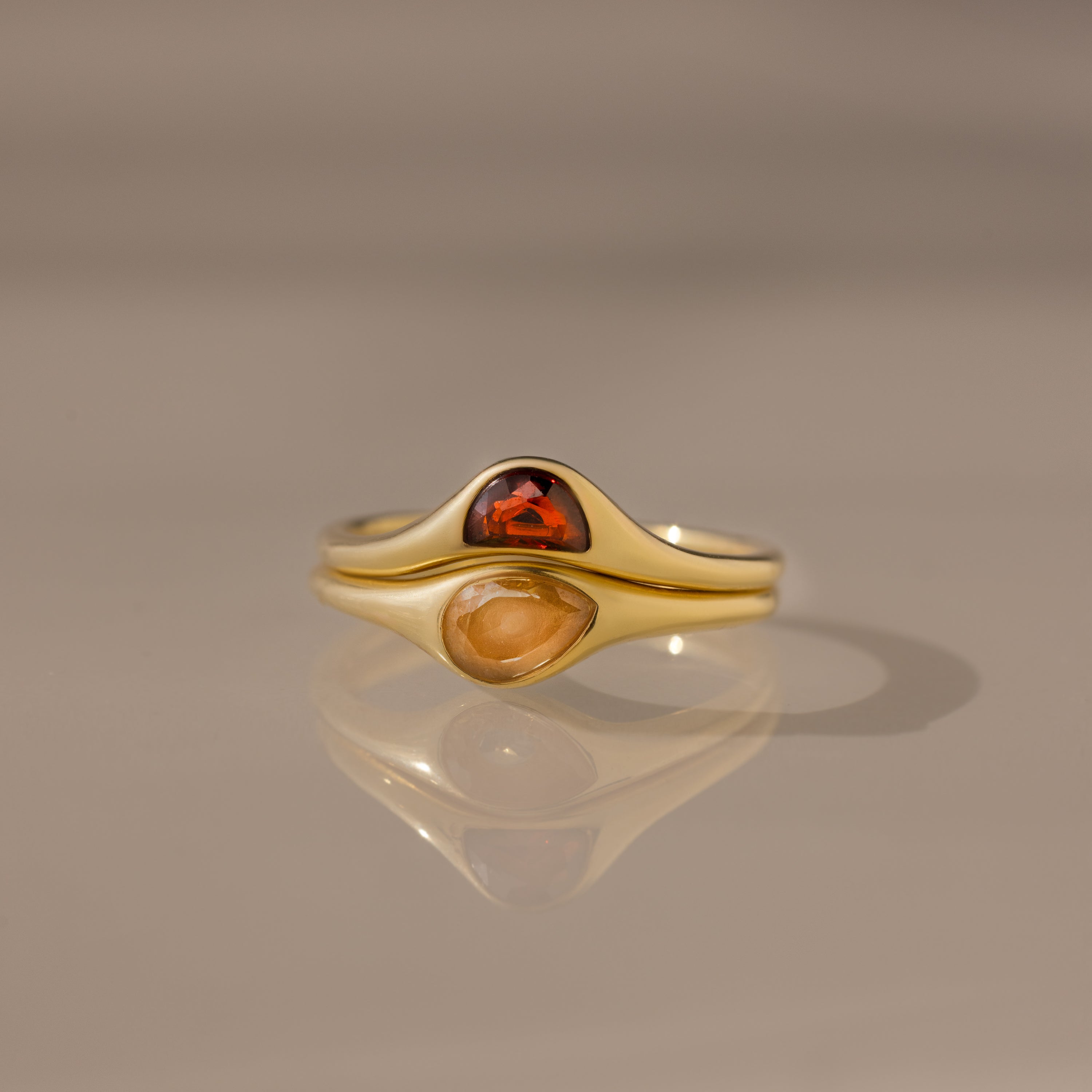 The Sculpted Toi et Moi Ring Set in 18K Gold features two gold rings with pear-shaped red and yellow gemstones, elegantly stacked on a smooth reflective surface.