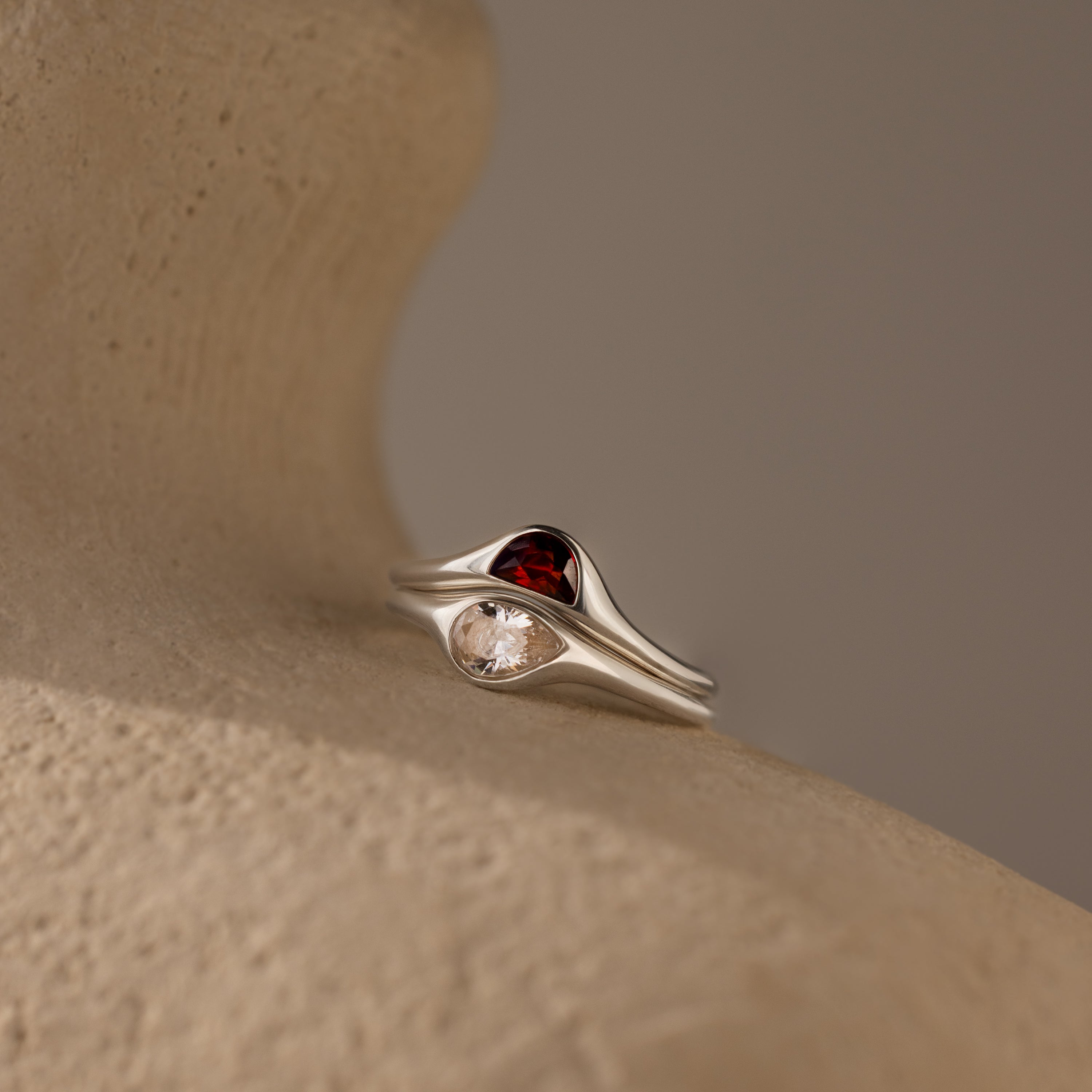 A Sculpted Toi et Moi Ring Set with one red and one clear gemstone sits on a textured beige surface.