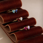 The Love Toi et Moi Ring in 18K Gold features four gold stacking rings with colorful heart-shaped gemstones, displayed on a brown velvet holder—perfect as a modern statement piece or unique gemstone ring.