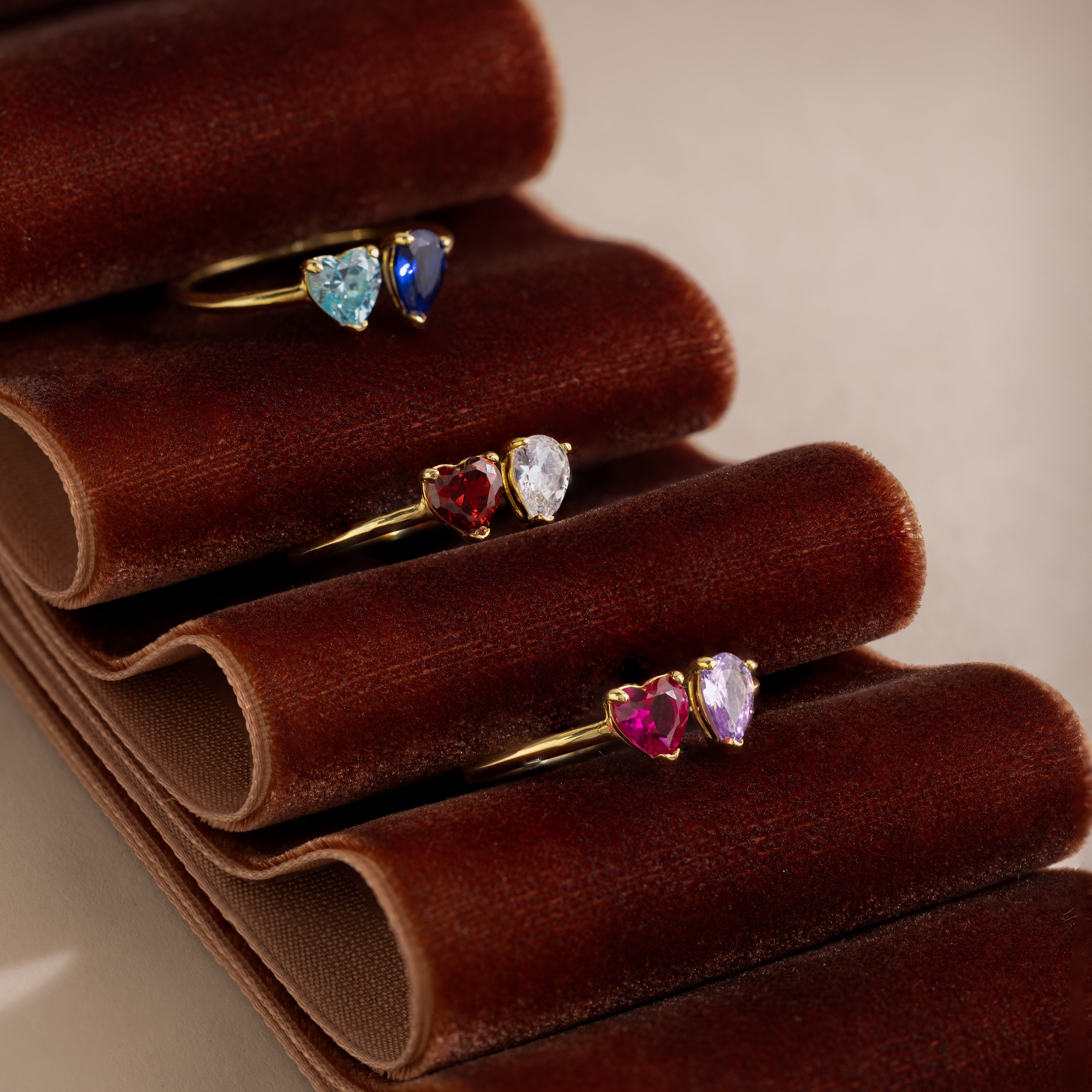 The Love Toi et Moi Ring in 18K Gold features four gold stacking rings with colorful heart-shaped gemstones, displayed on a brown velvet holder—perfect as a modern statement piece or unique gemstone ring.