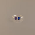 The Love Toi et Moi Ring in Silver features two heart-shaped gemstones—one purple and one blue—set side by side and shown against a plain neutral background.