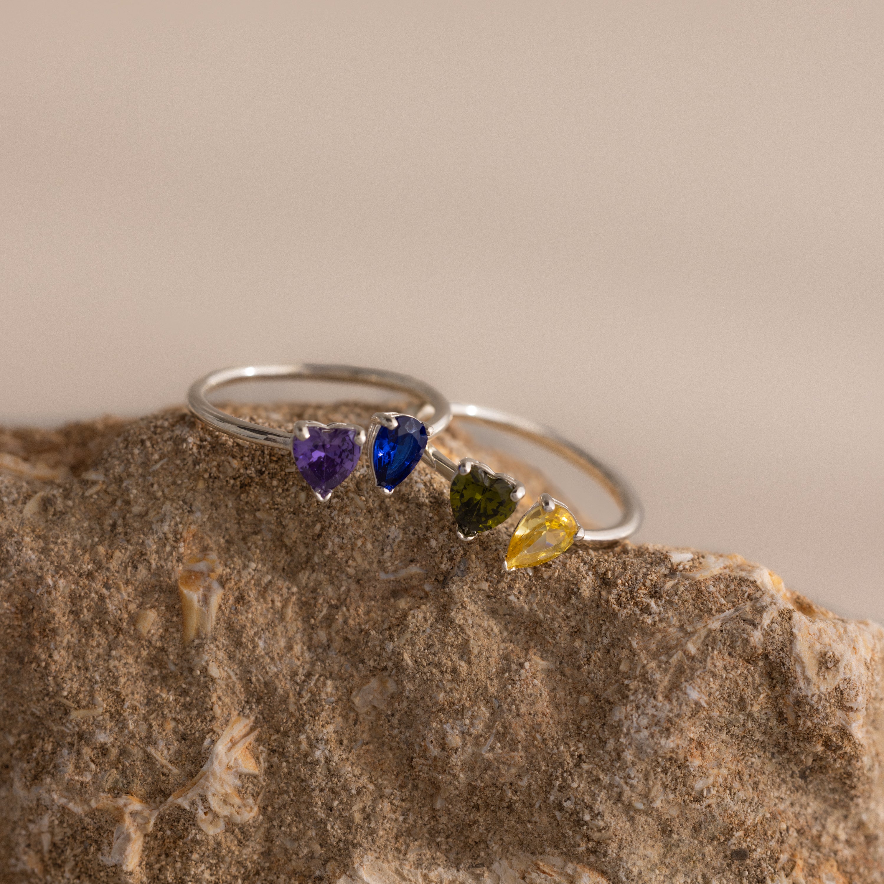 The Love Toi et Moi Ring in Silver features heart-shaped purple, blue, green, and yellow gemstones stacked together and displayed on a textured rock.