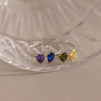 The Love Toi et Moi Ring features four silver stacking bands, each with a heart-shaped gemstone in purple, blue, green, and yellow, displayed on a glass surface.