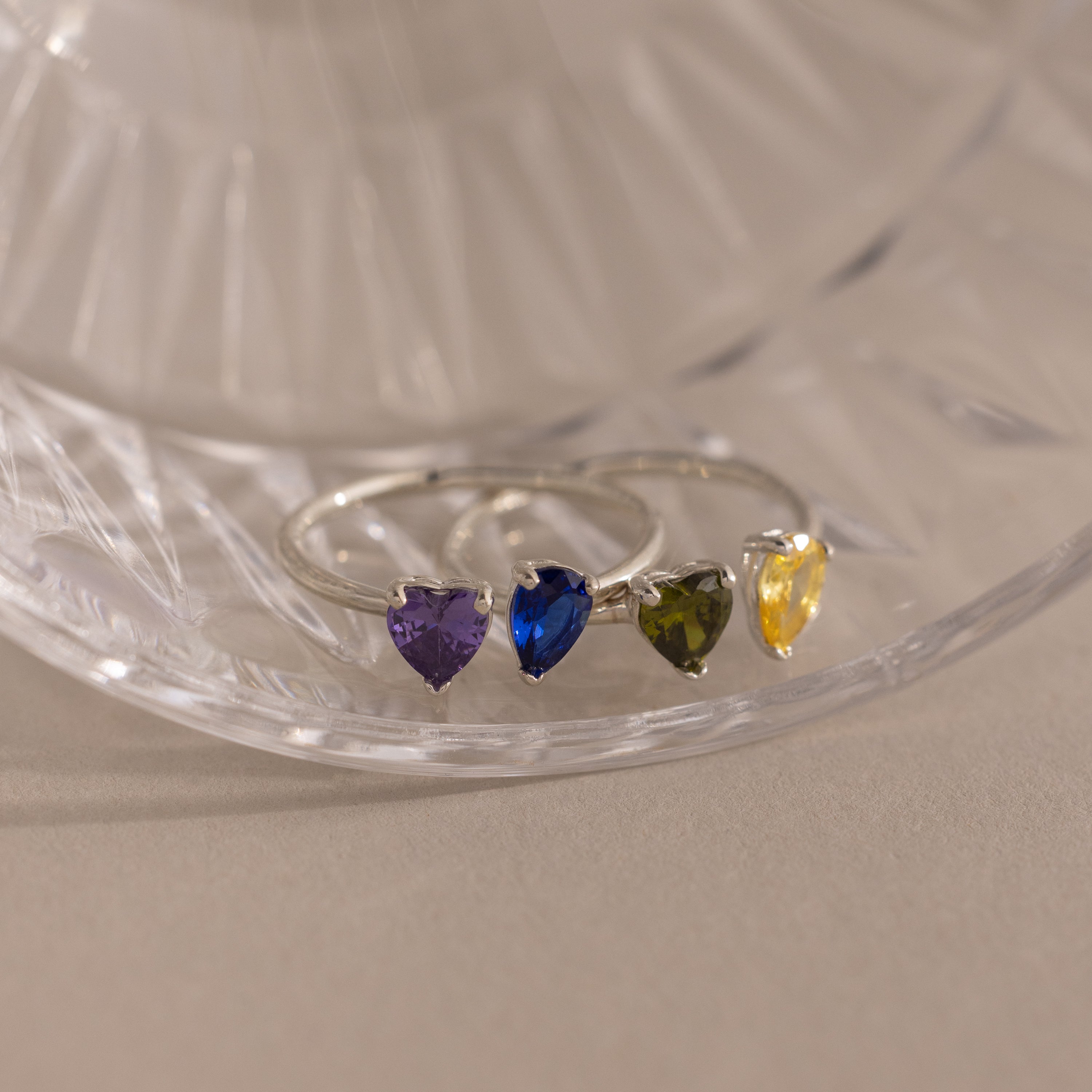 The Love Toi et Moi Ring features four silver stacking bands, each with a heart-shaped gemstone in purple, blue, green, and yellow, displayed on a glass surface.