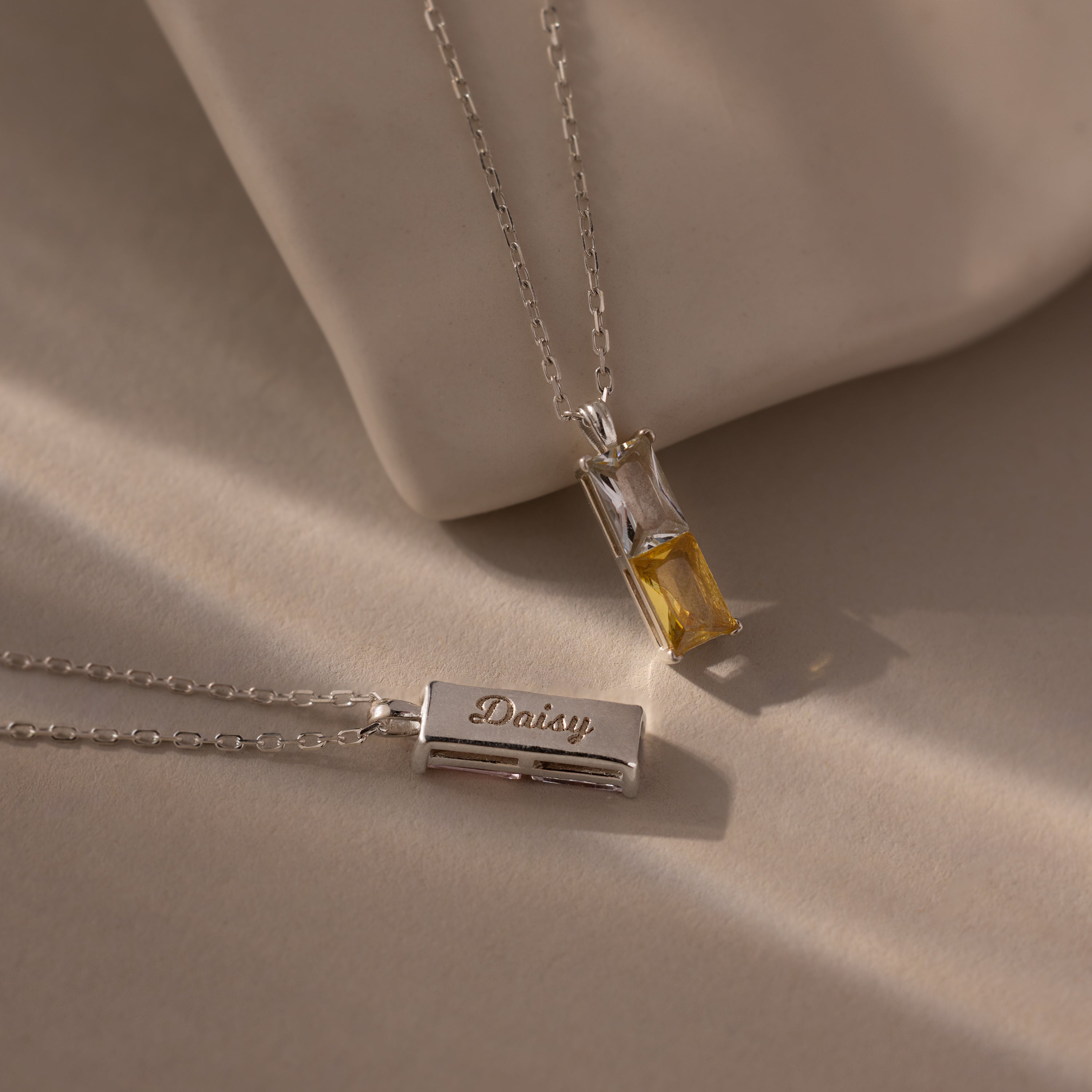 The Duo Birthstone Name Necklace set includes two silver necklaces: one with a yellow gemstone, the other with a rectangular pendant engraved with Daisy—an ideal personalized gift featuring custom name engraving.
