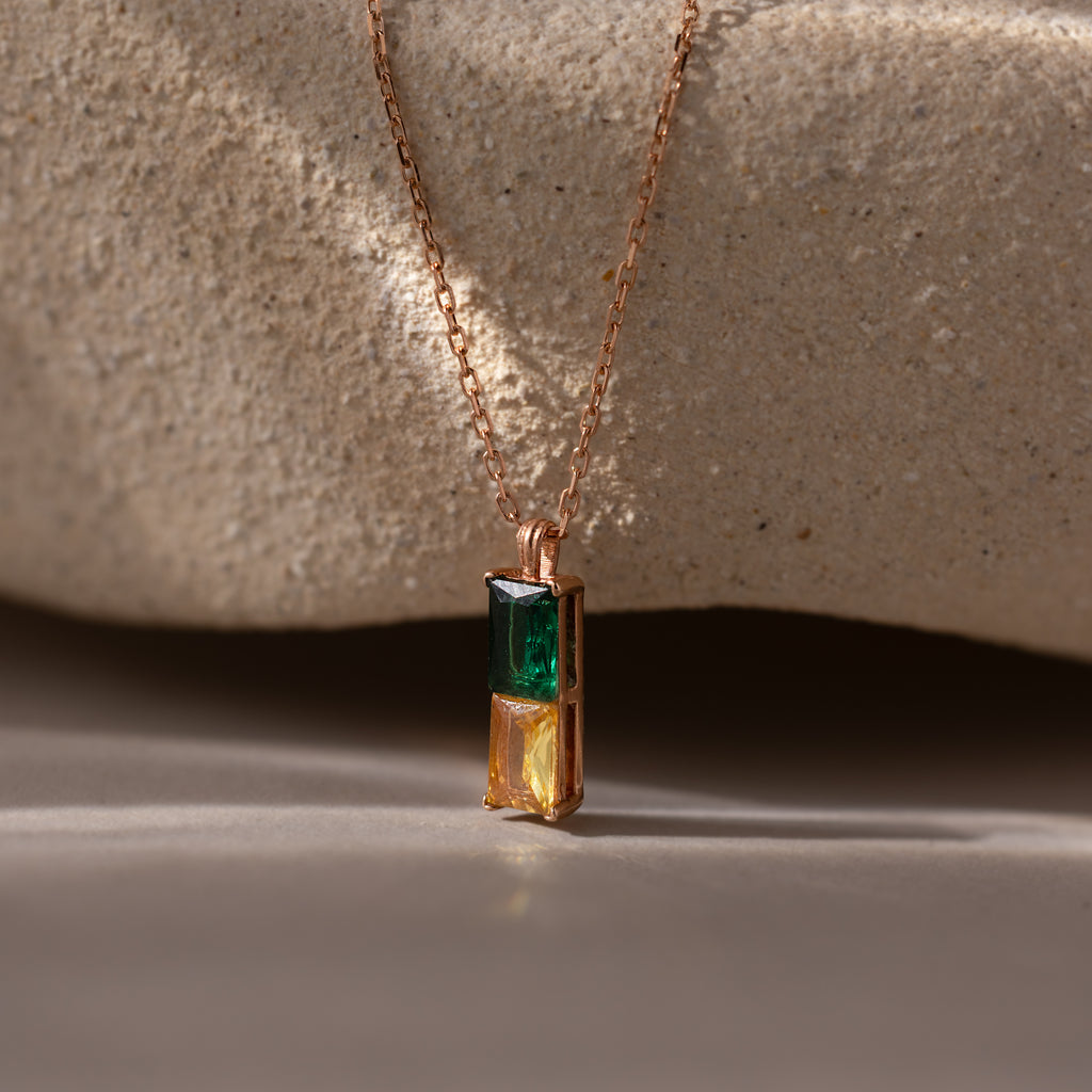 The Duo Birthstone Name Necklace in Rose Gold features elegant green and yellow rectangular stones, set against a textured beige stone background—a stylish personalized jewelry piece perfect for custom name engraving.