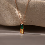 The Duo Birthstone Name Necklace in Rose Gold features elegant green and yellow rectangular stones, set against a textured beige stone background—a stylish personalized jewelry piece perfect for custom name engraving.