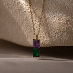 The Duo Birthstone Name Necklace in 18K Gold features purple and green rectangular gemstones, displayed on textured beige stone. Personalize this stunning keepsake with a custom name engraving for a unique birthstone necklace.
