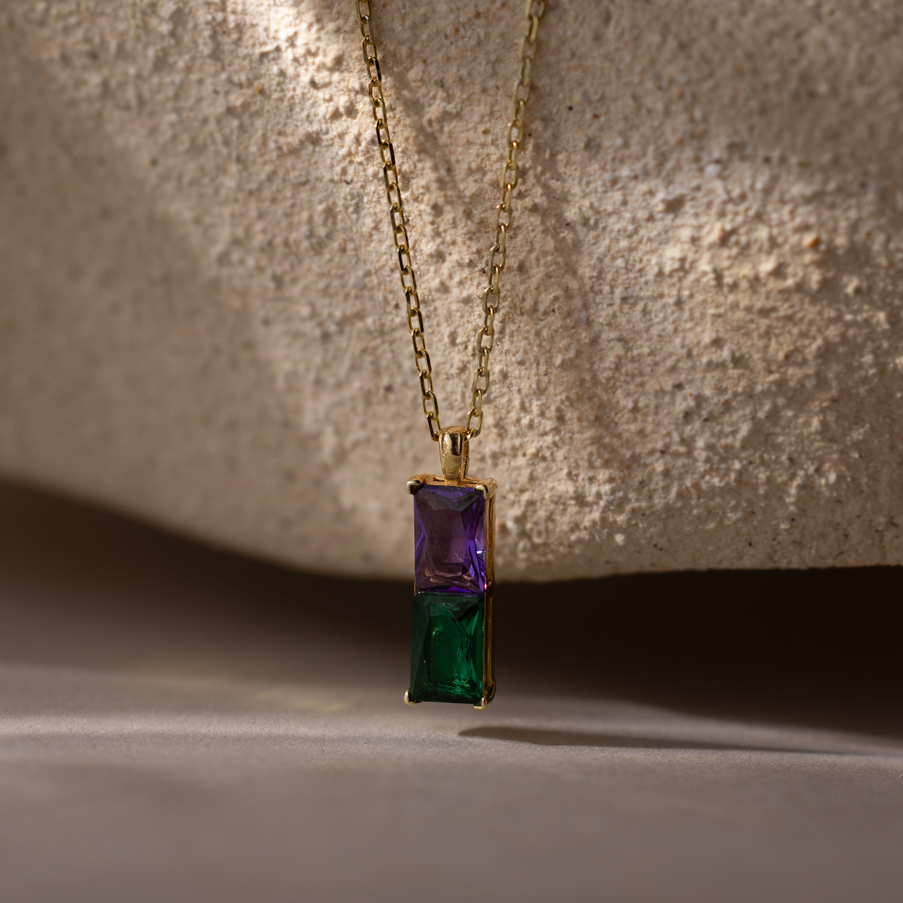 The Duo Birthstone Name Necklace in 18K Gold features purple and green rectangular gemstones, displayed on textured beige stone. Personalize this stunning keepsake with a custom name engraving for a unique birthstone necklace.