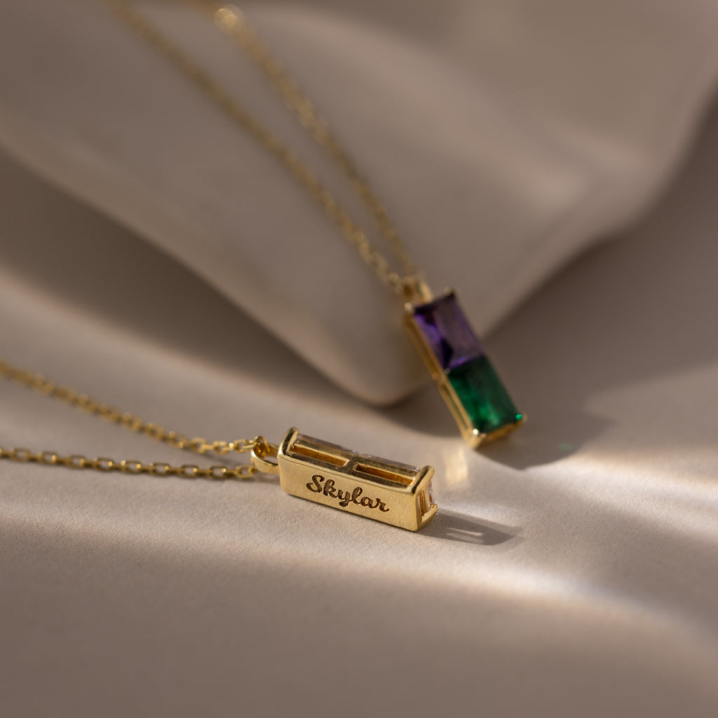 The Duo Birthstone Name Necklace in 18K Gold features two gold pendants on chains, each with rectangular green and purple gemstones—one personalized with the name Skylar for a unique keepsake.