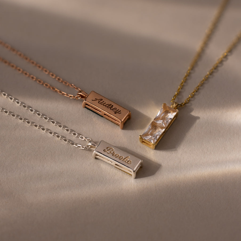 The Duo Birthstone Name Necklace in Rose Gold is displayed on a beige surface, featuring a rectangular gemstone pendant and slender bars personalized with engraved names.