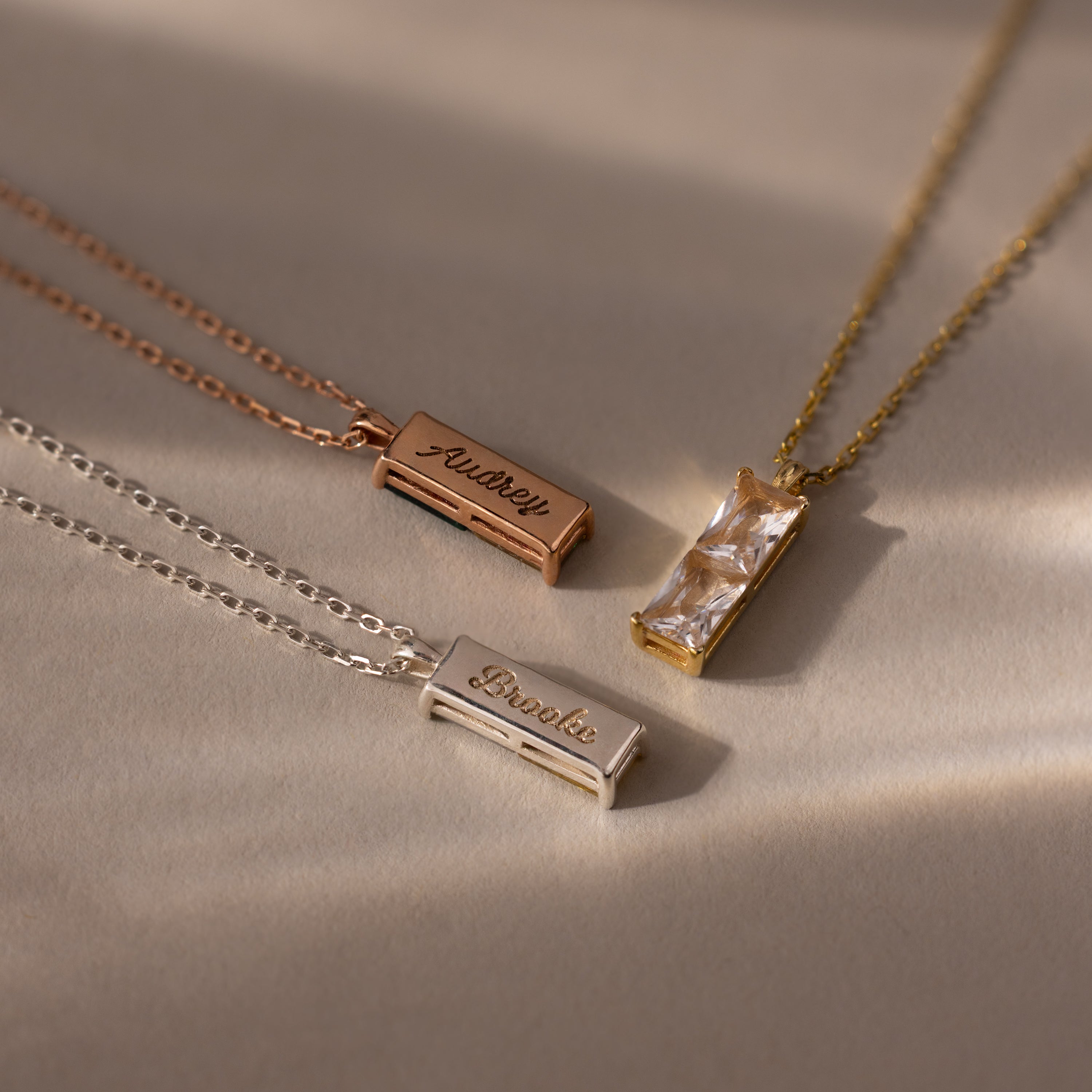 The Duo Birthstone Name Necklace in Rose Gold is displayed on a beige surface, featuring a rectangular gemstone pendant and slender bars personalized with engraved names.