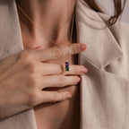 A woman in a beige blazer holds the Duo Birthstone Name Necklace, a gold chain featuring a rectangular pendant with purple and green gemstones—an ideal personalized keepsake.