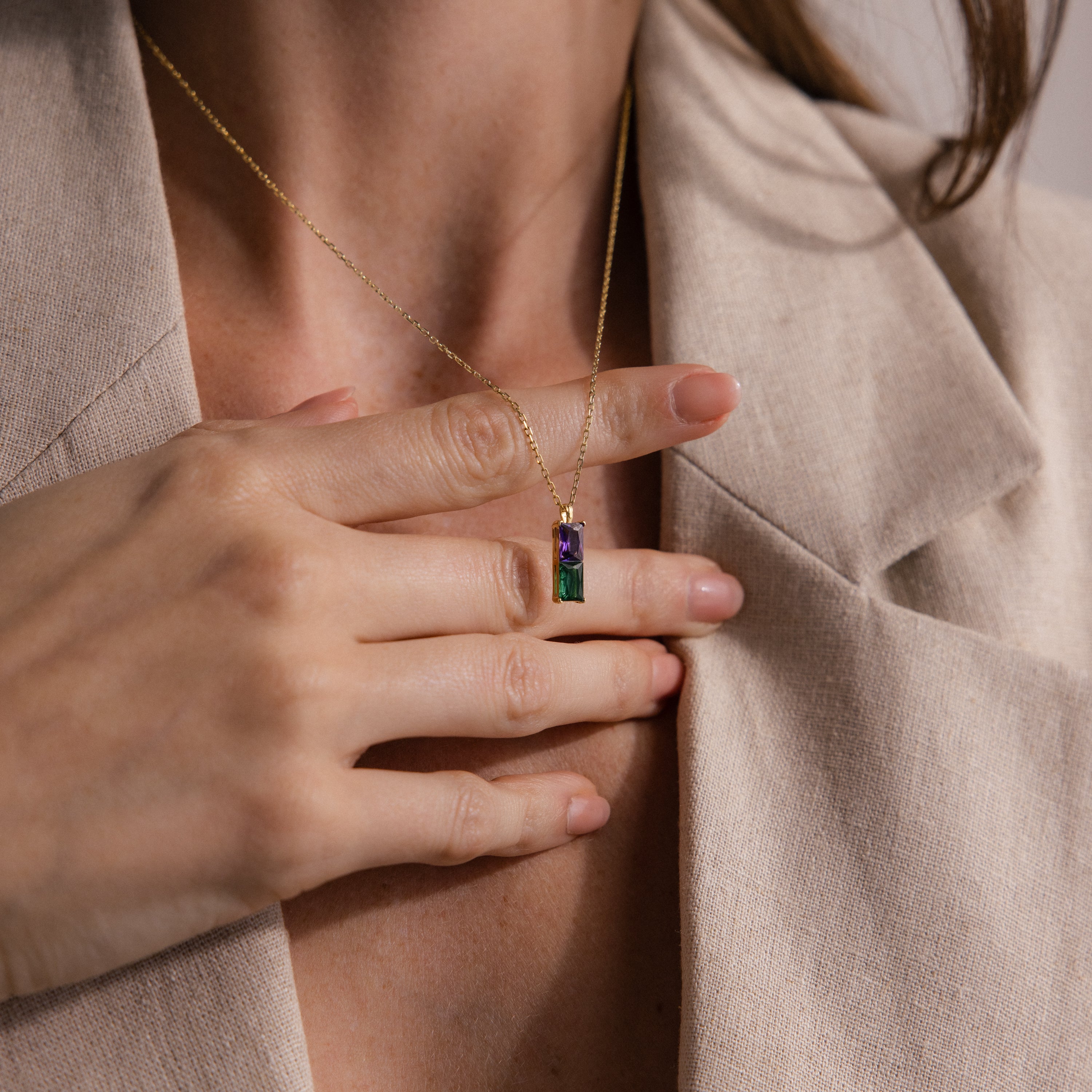 A woman in a beige blazer holds the Duo Birthstone Name Necklace, a gold chain featuring a rectangular pendant with purple and green gemstones—an ideal personalized keepsake.