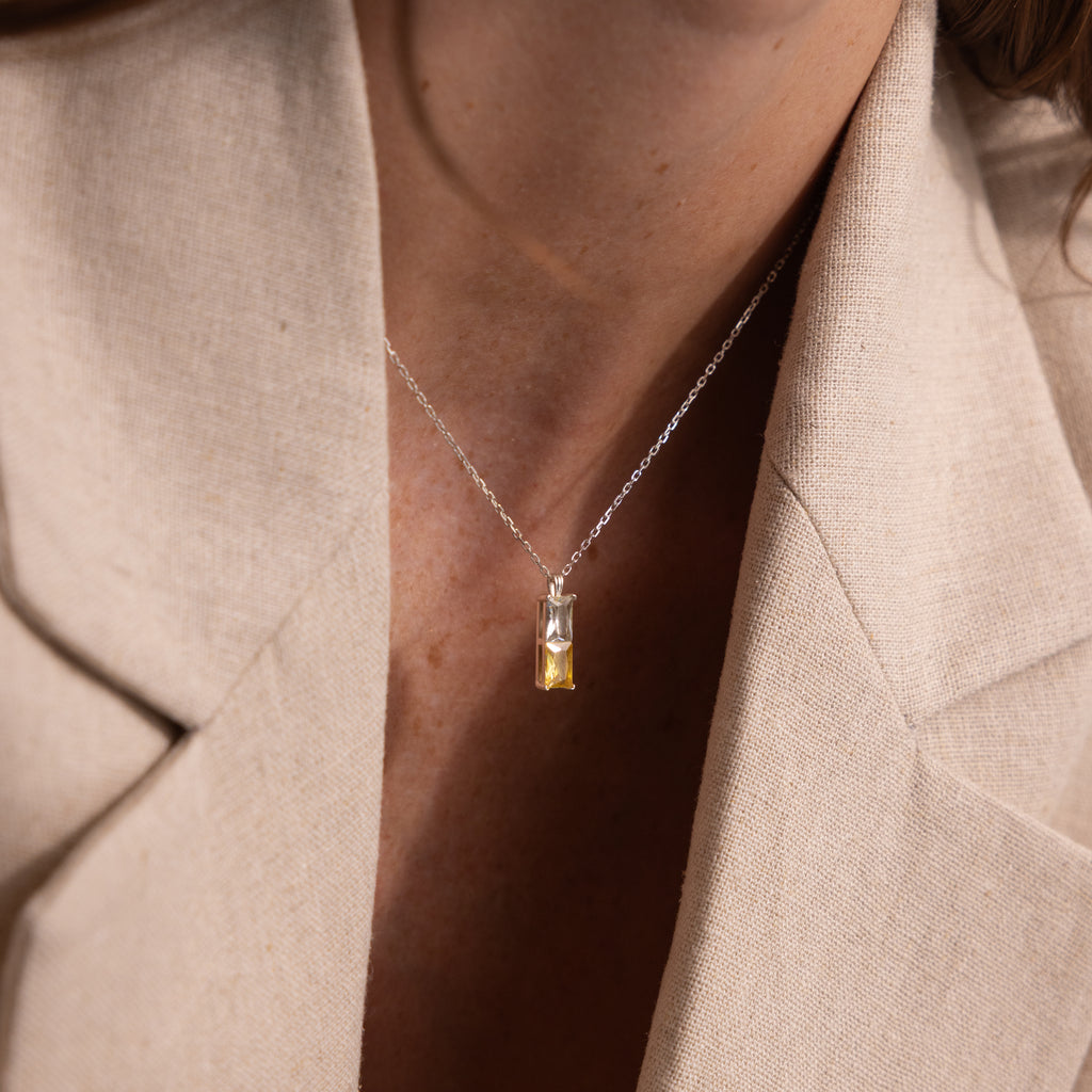 A woman wears a beige blazer and the Duo Birthstone Name Necklace in Sterling Silver, featuring a rectangular yellow gemstone pendant for a personalized touch.