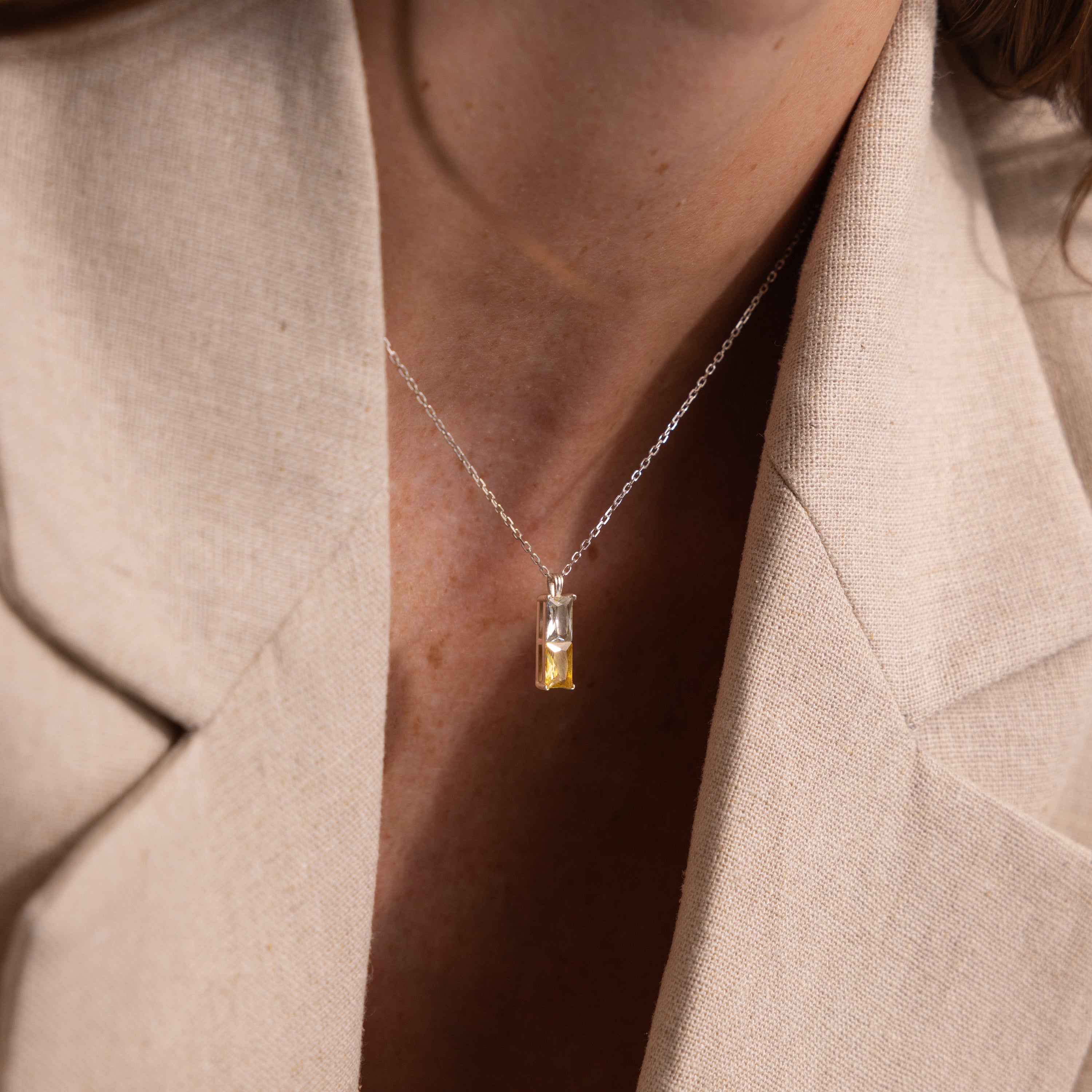 A woman wears a beige blazer and the Duo Birthstone Name Necklace in Sterling Silver, featuring a rectangular yellow gemstone pendant for a personalized touch.