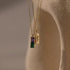 The Duo Birthstone Name Necklace in 18K Gold features two necklaces: a personalized birthstone name necklace with colorful stones and another with an engraved rectangular pendant, creating a unique keepsake of self love.