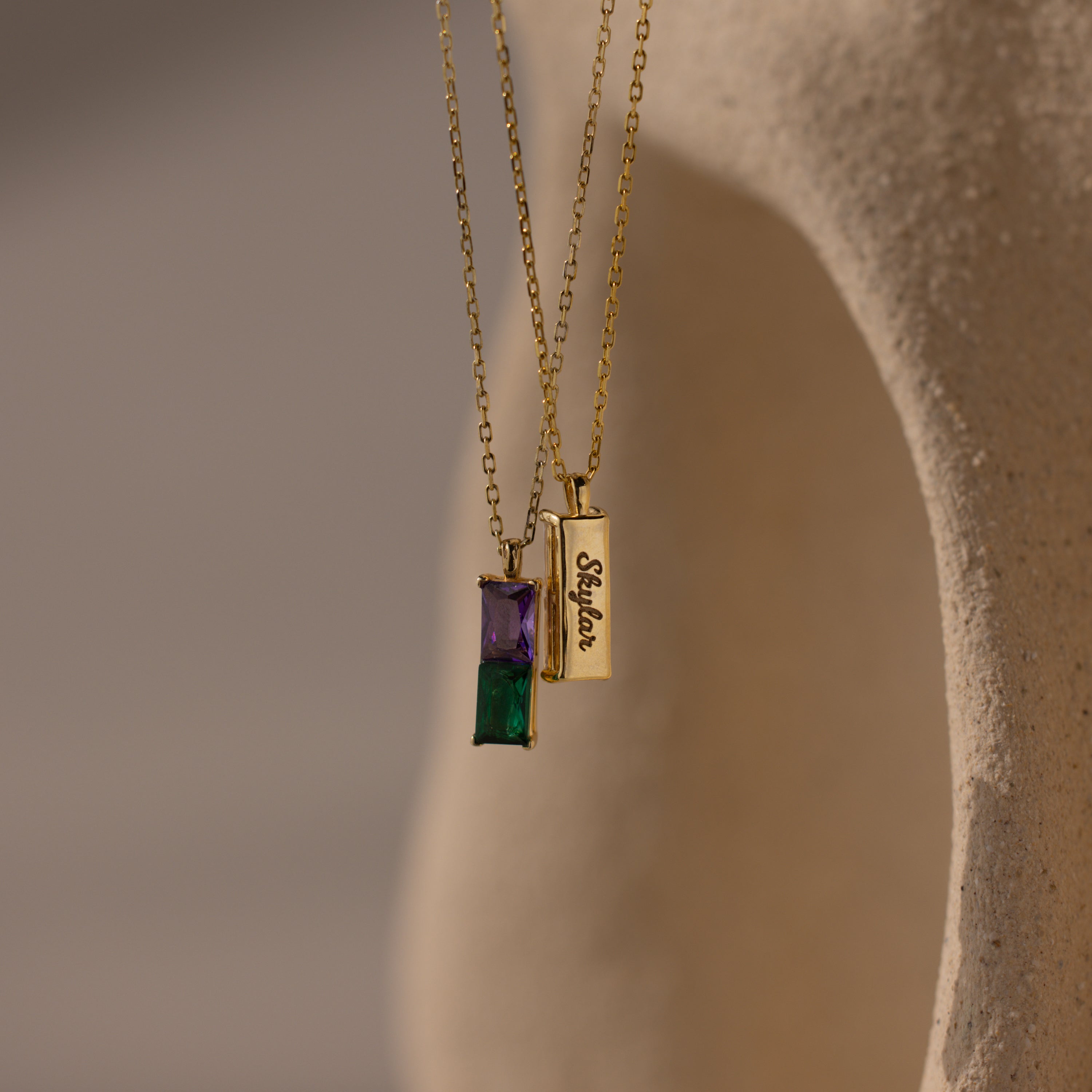 Two gold necklaces—a Duo Birthstone Name Necklace with purple and green stones, and a rectangular self-love pendant—are displayed on a textured background, offering a perfect personalized keepsake.