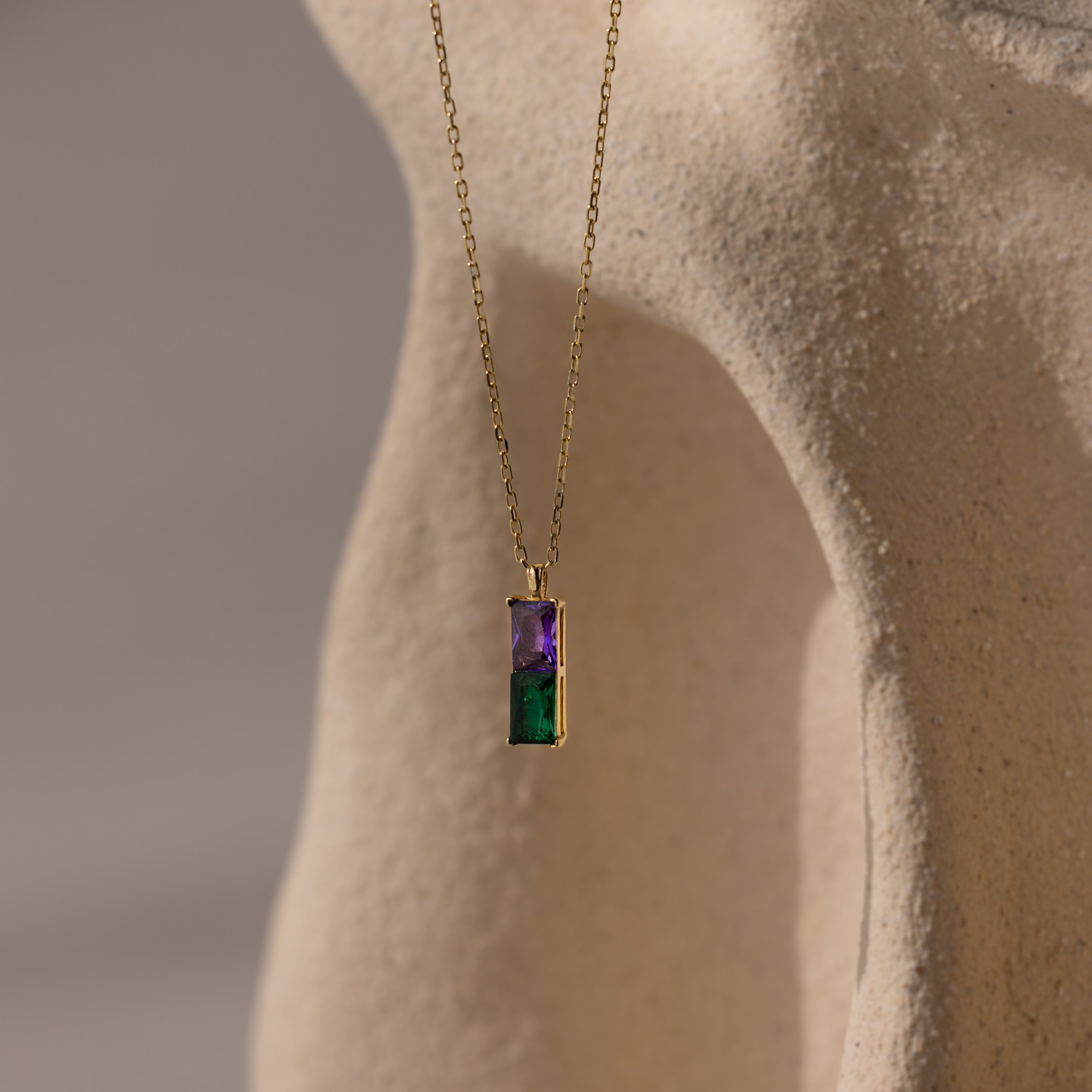 Duo Birthstone Name Necklace in 18K Gold with purple and green rectangular stones, displayed on a textured beige background—this personalized piece is a stunning keepsake.