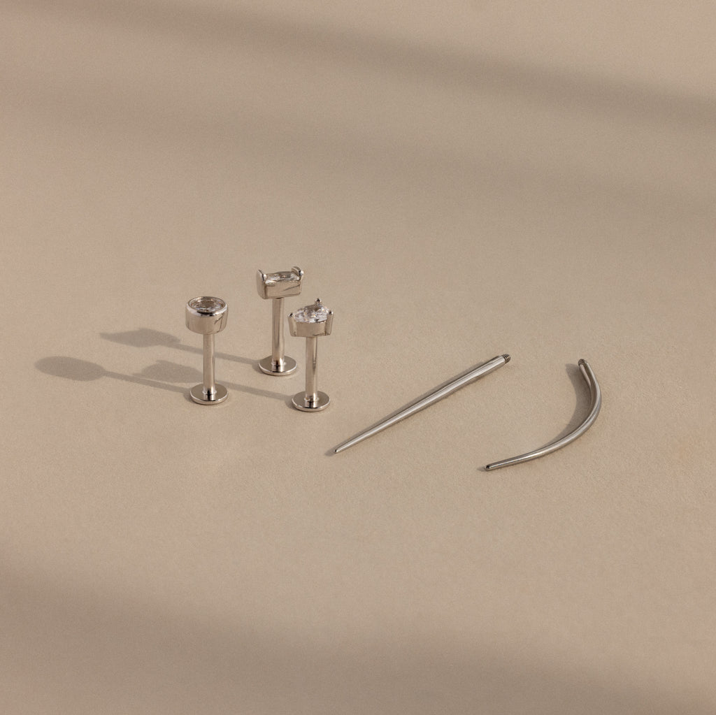 A Flatback Applicator Tool is shown on a beige surface beside three silver stud earrings with clear gemstones and two curved silver barbell earrings.