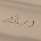 A Flatback Applicator Tool is shown on a beige surface beside three silver stud earrings with clear gemstones and two curved silver barbell earrings.