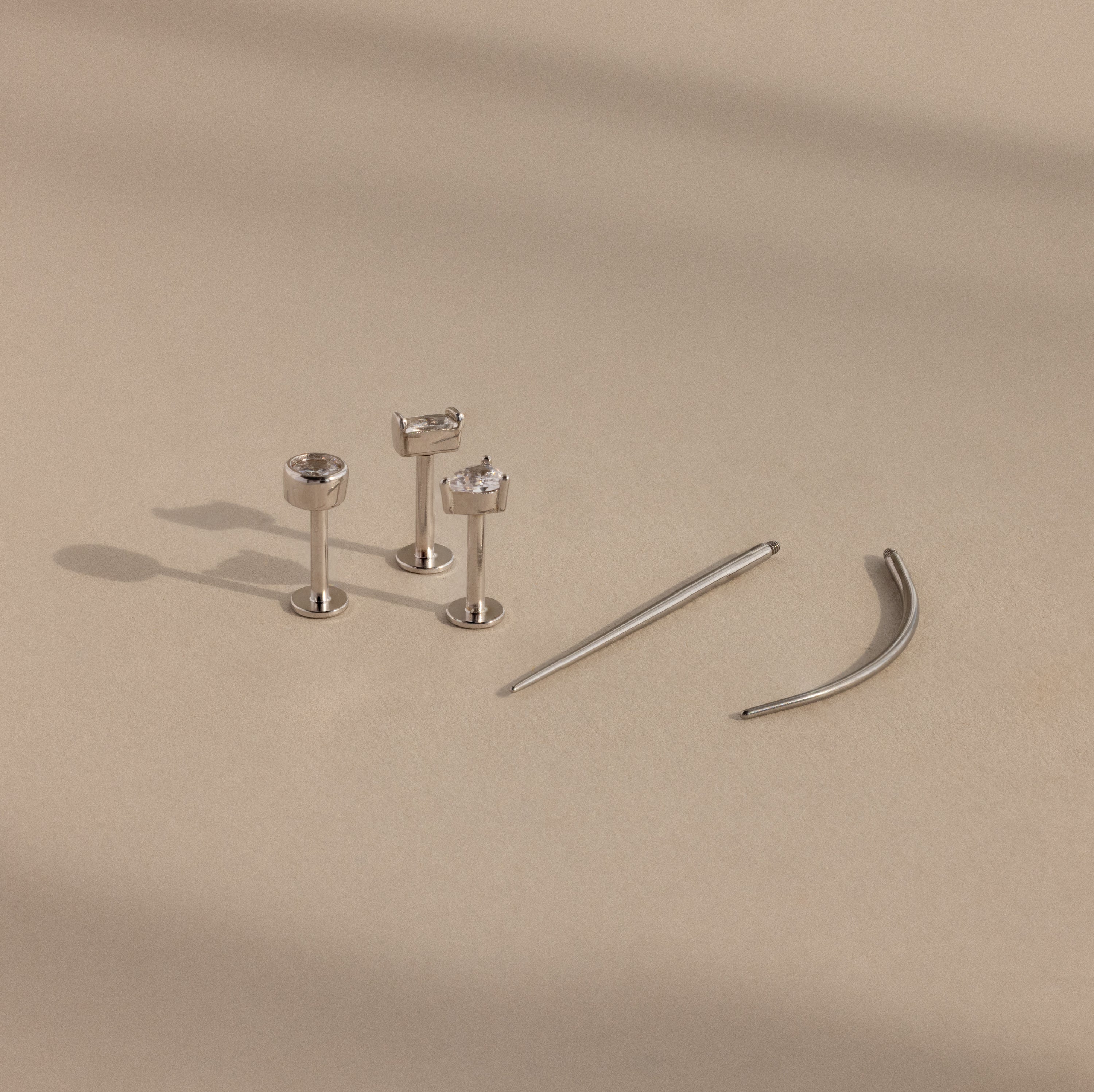 A Flatback Applicator Tool is shown on a beige surface beside three silver stud earrings with clear gemstones and two curved silver barbell earrings.