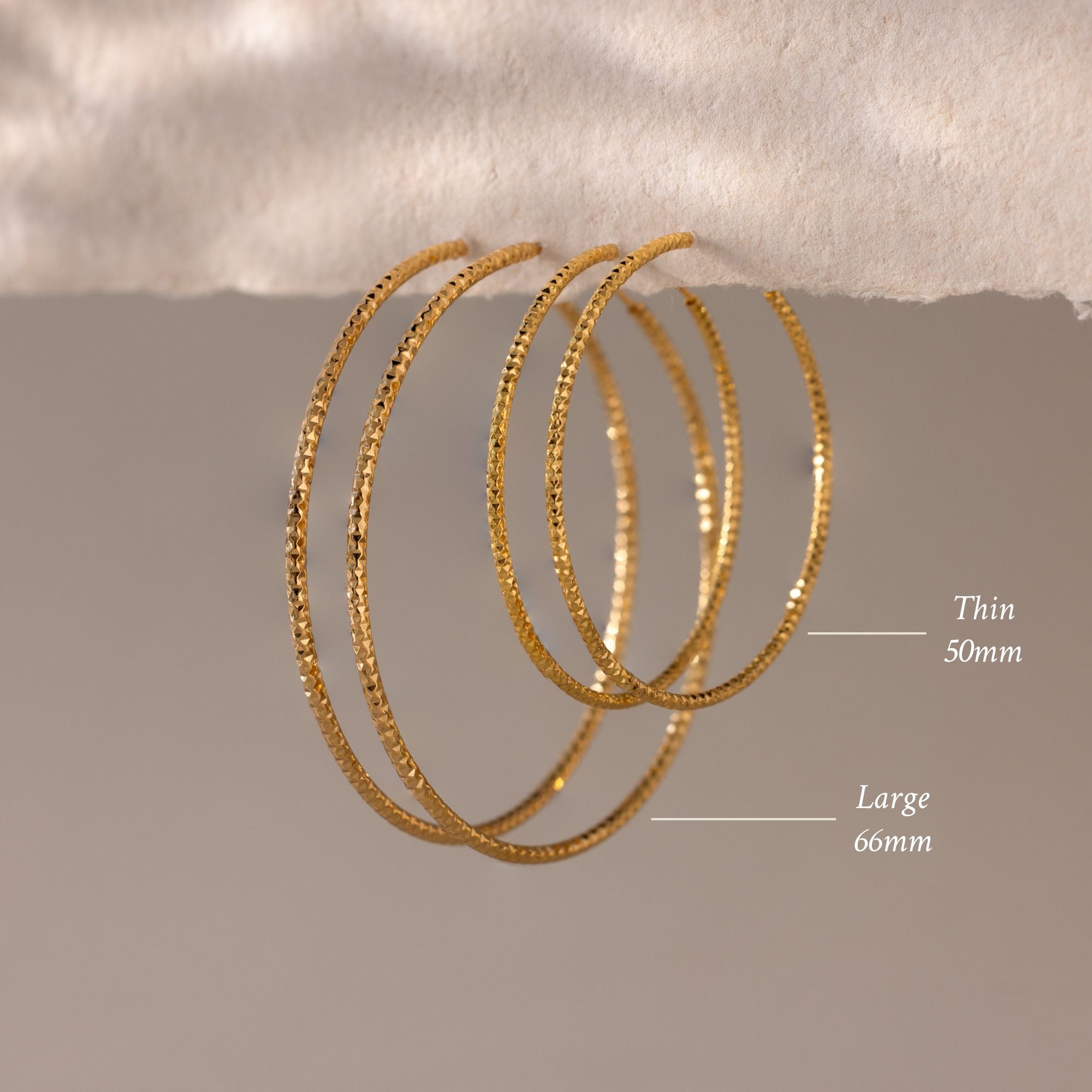 A close-up image of four gold textured hoop earrings hanging elegantly from a soft, torn-edge piece of paper. The hoops have a subtle hammered finish that reflects the light, creating a shimmering effect against the neutral beige background.